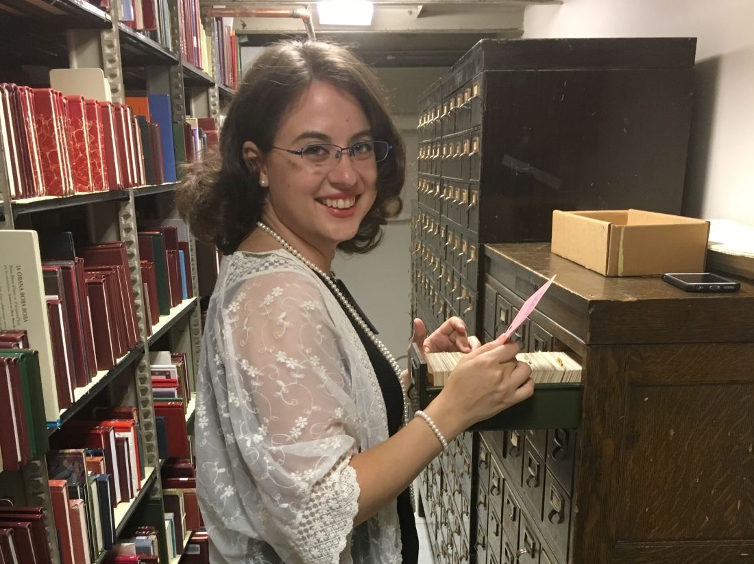 Anna Groves smiles at the camera while flipping through index cards in a wooden card catalog drawer, standing in an archive room with shelves of red and green bound volumes at the Library of Congress.