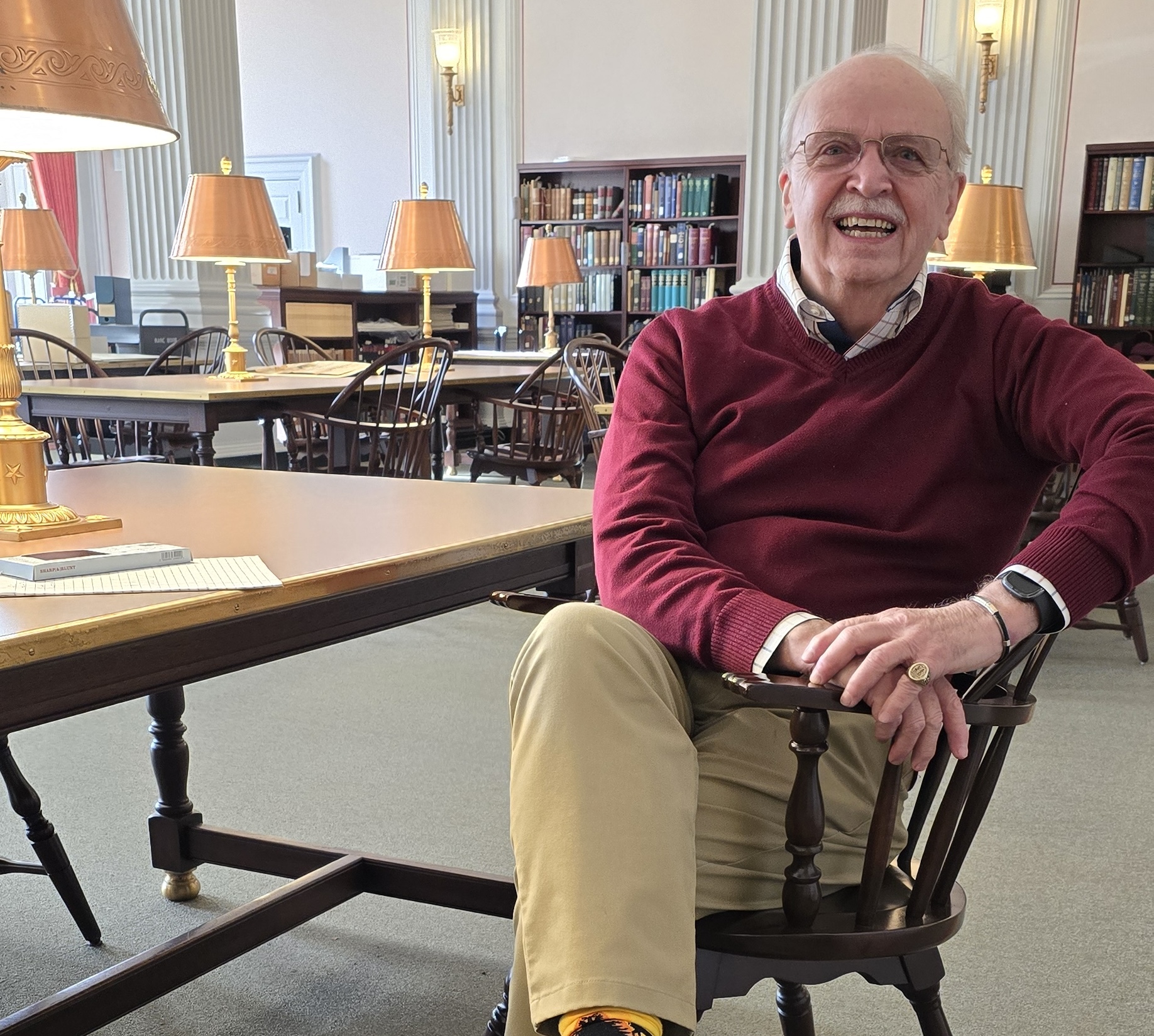 Brent Kendrick sits smiling in a wooden chair in a reading room at the Library of Congress, wearing a maroon sweater, with long wooden tables, brass lamps, and bookshelves filled with volumes in the background.