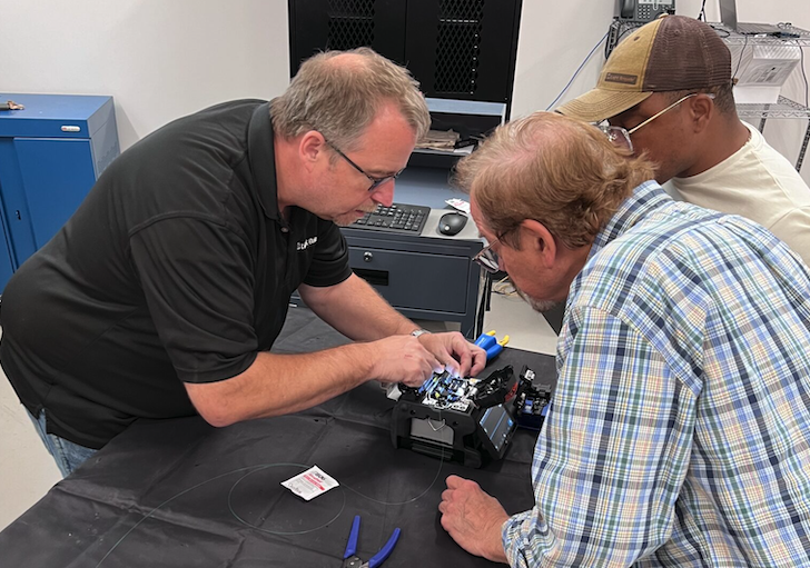 People participating in hands-on fiber optic splicing training, gathered around a fiber splicing machine in a technical lab setting.