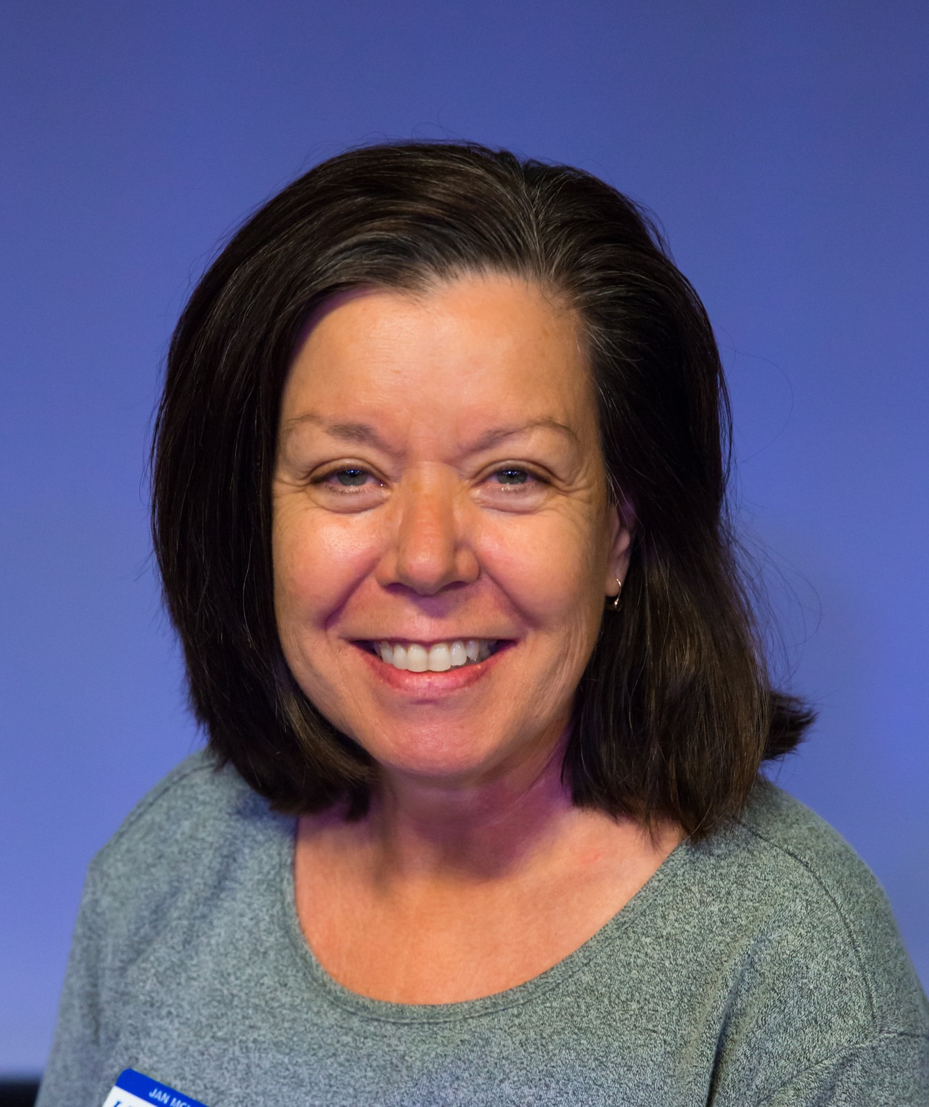 Headshot of a Jan McLaughlin with shoulder-length dark hair wearing a light gray top against a blue background.