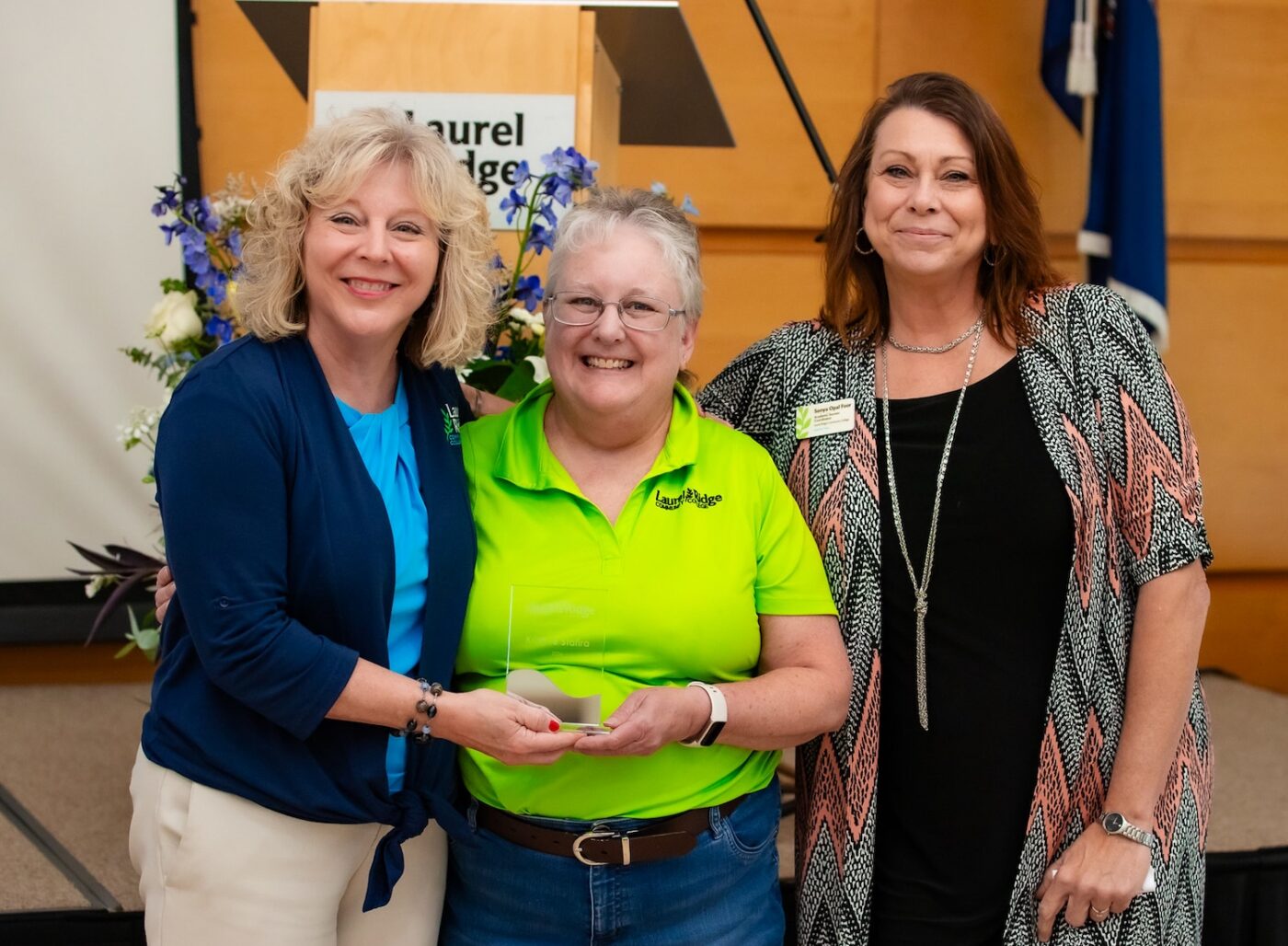Three women stand together at a Laurel Ridge Community College event, smiling for the camera. The woman in the center wears a bright green Laurel Ridge polo shirt and holds a glass award, flanked by two women in professional attire with their arms around her. A podium with the Laurel Ridge logo and a floral arrangement are visible in the background.