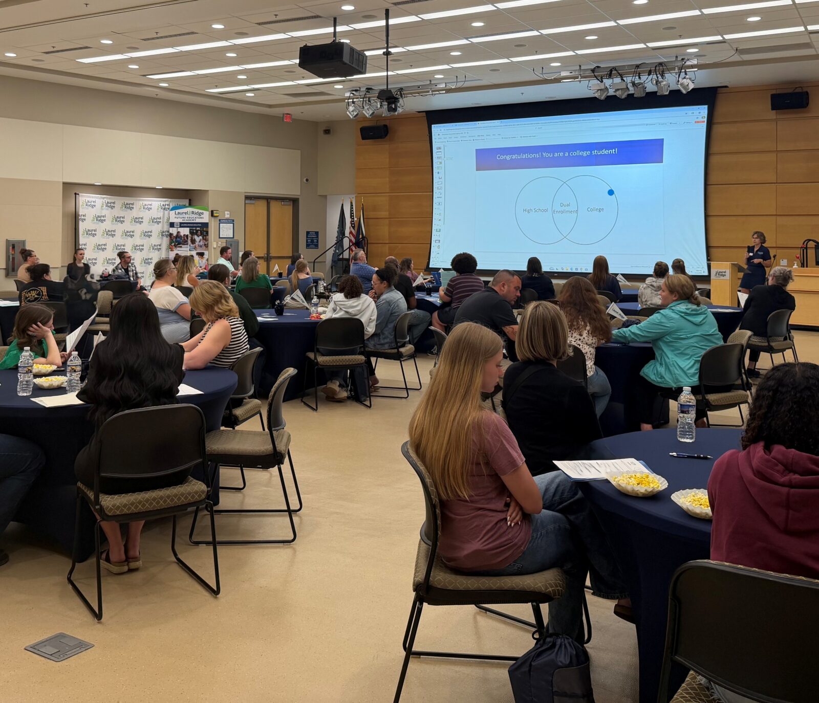 A large room full of attendees seated at round tables with navy tablecloths, watching a presentation at a Laurel Ridge Community College event. On the projector screen, a slide reads "Congratulations! You are a college student!" with a Venn diagram showing "High School," "Dual Enrollment," and "College" overlapping circles. A woman, Lori K., stands at a podium near the screen presenting. Laurel Ridge and Future Educators Academy banners are visible in the background, along with American and Virginia state flags. Attendees have papers, water bottles, and popcorn on their tables.