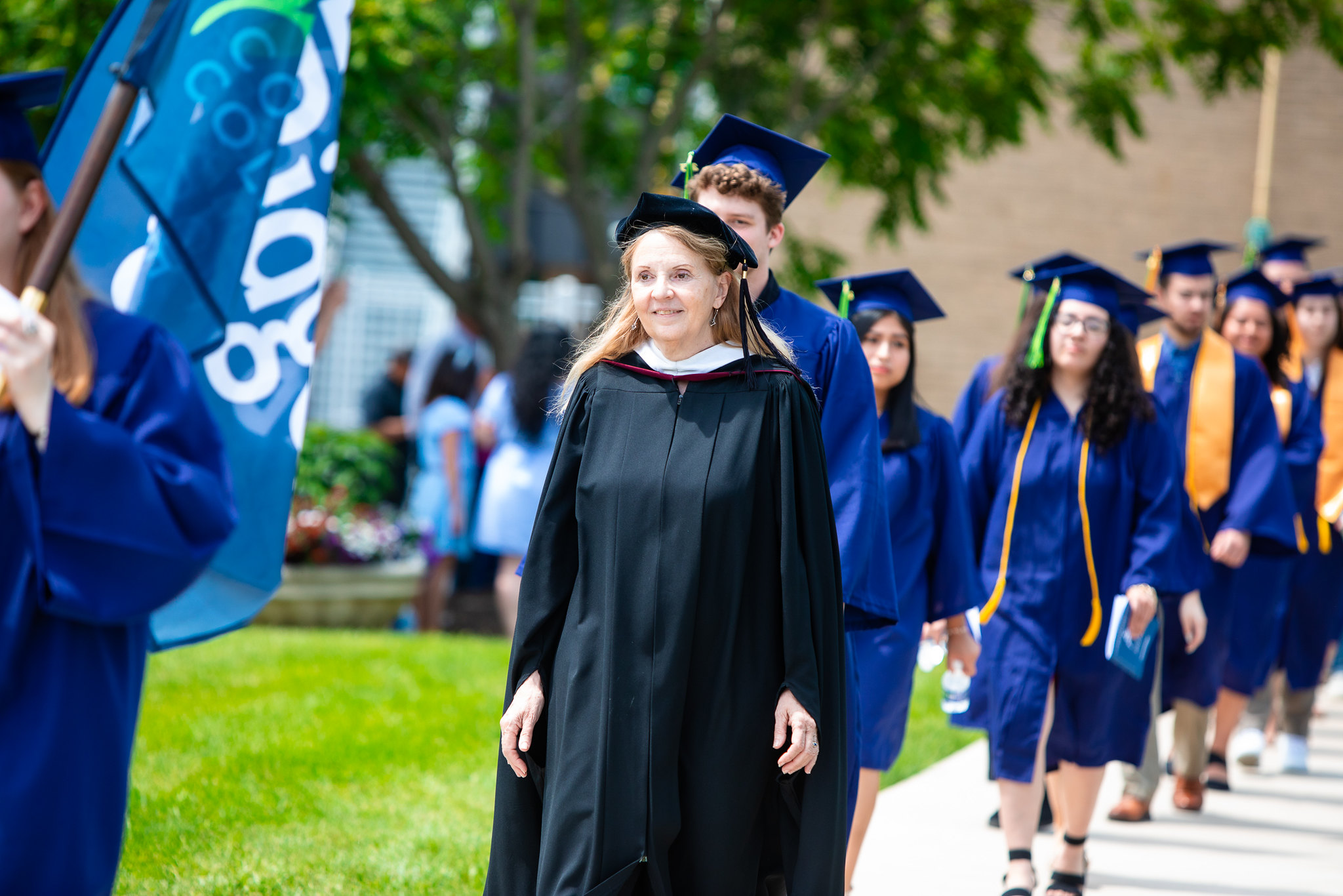 Nicole Martine in academic regalia leading a commencement procession with students in blue caps and gowns walking on a campus sidewalk.