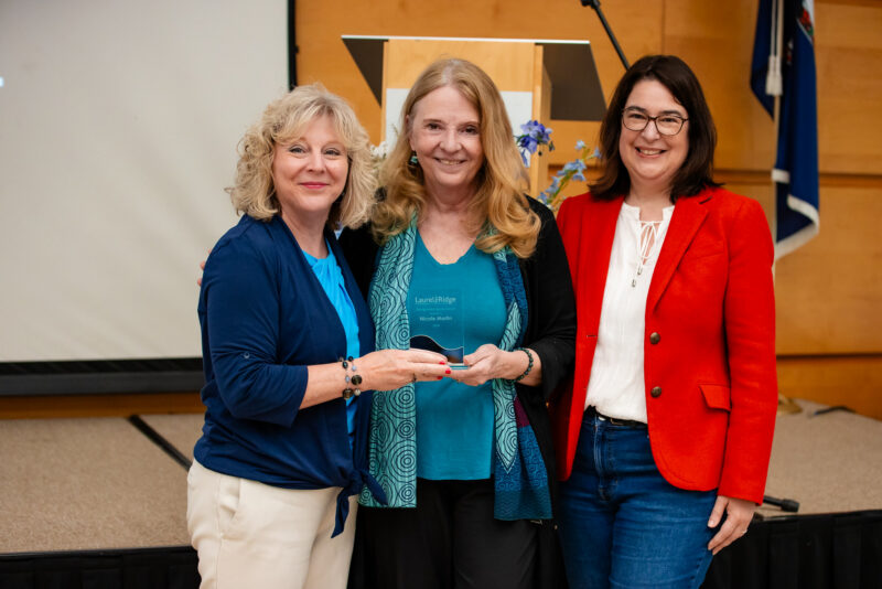 Three people standing together on a stage during an award presentation, with the center person holding a glass award and a podium in the background.