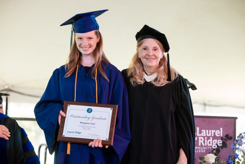 Graduate in blue cap and gown holding an award plaque while standing beside a faculty Nicole Martin member in academic regalia at a commencement ceremony.