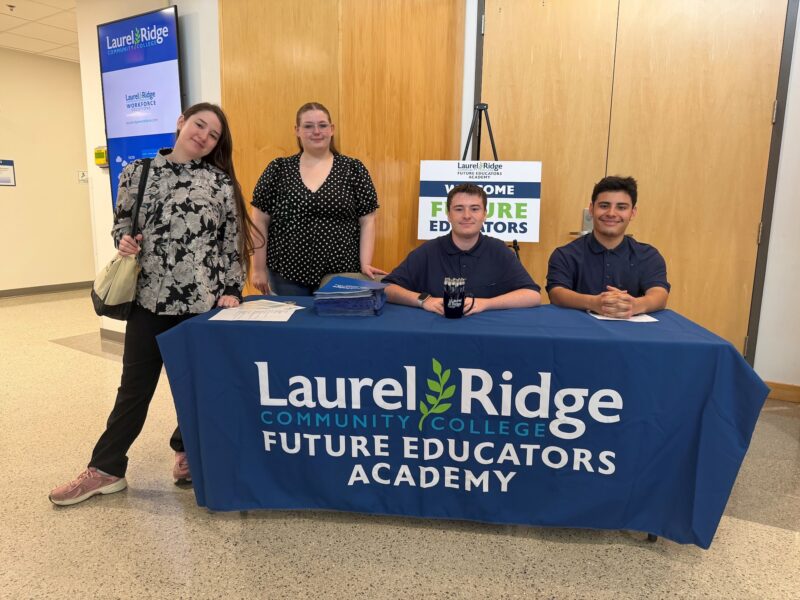 Four smiling people pose at a Laurel Ridge Community College Future Educators Academy welcome table in a hallway. Two young women stand on the left side of the table, one in a gray floral top and one in a black polka-dot blouse, while two young men in matching navy polo shirts sit behind the table. The table is covered with a navy blue tablecloth reading "Laurel Ridge Community College Future Educators Academy," and holds stacks of blue folders, a mug of pens, and papers. A sign behind them reads "Welcome Future Educators," and a Laurel Ridge digital display is visible in the background.