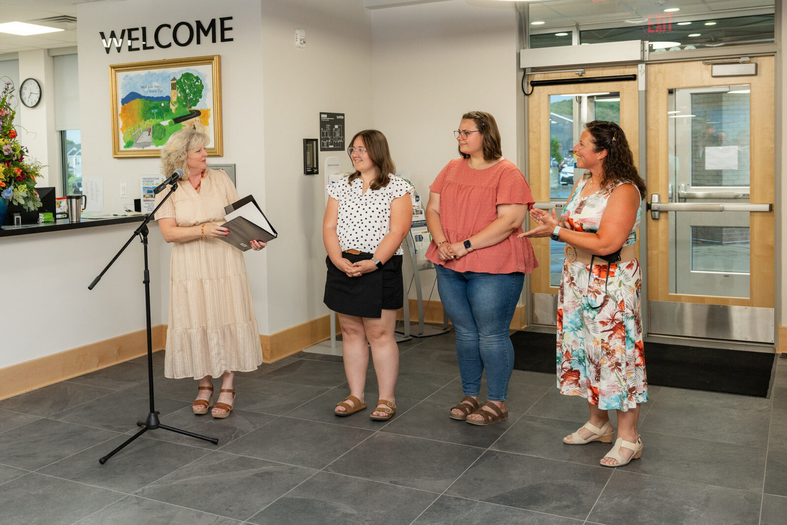Four women stand in a lobby beneath a "Welcome" sign at a Laurel Ridge Community College campus, near a colorful painted mural and a floral arrangement. A woman in a cream dress speaks into a microphone stand while holding a folder, addressing two young women, one in a black-and-white polka-dot top and black skirt, and the other in a coral top and jeans. A fourth woman in a floral skirt and white top gestures toward the two younger women. Glass entrance doors and a reception desk are visible in the background.