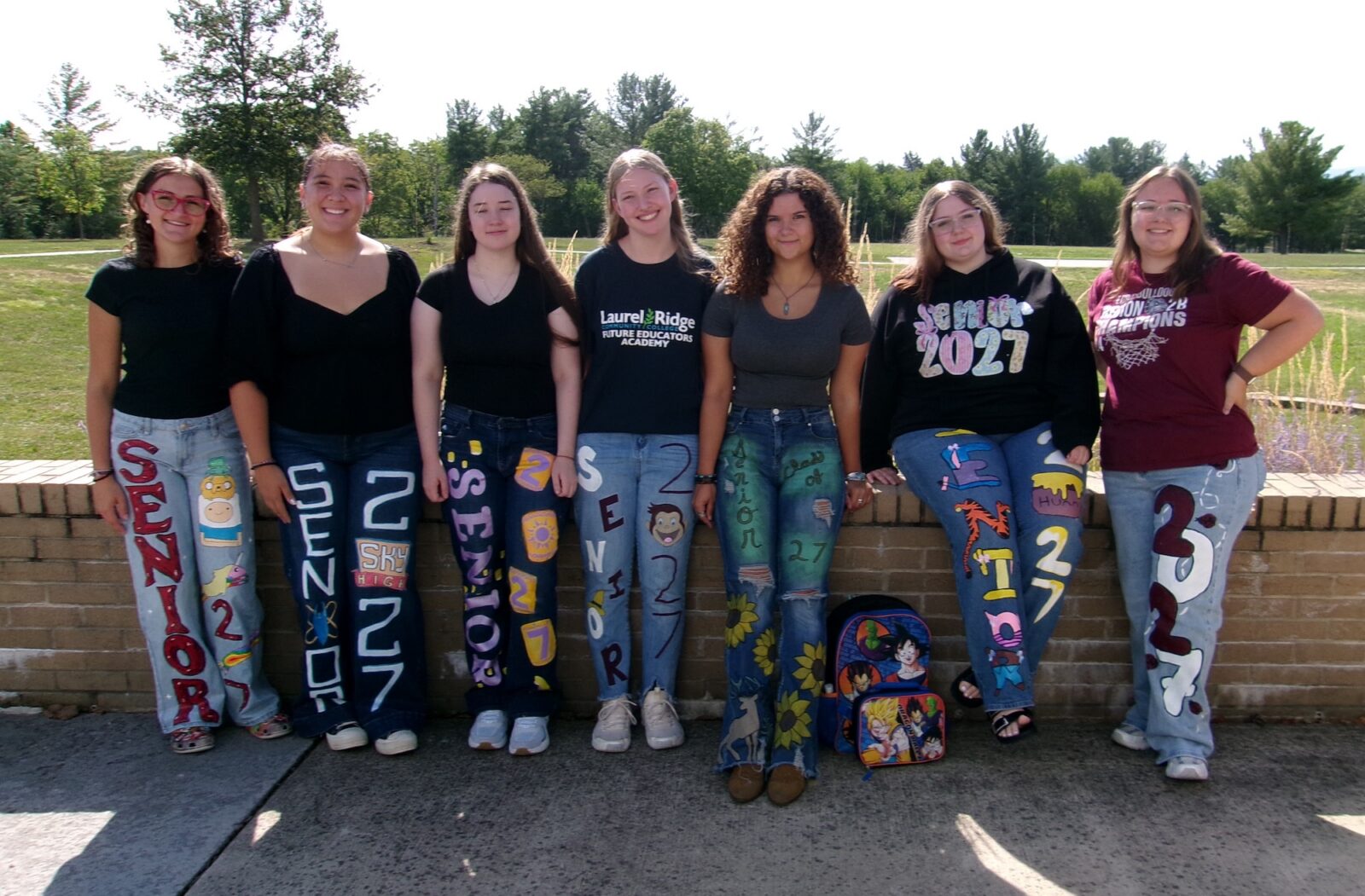Seven Future Educators Academy students lean against a brick wall outdoors, each wearing hand-painted "Senior 2027" designs on their jeans