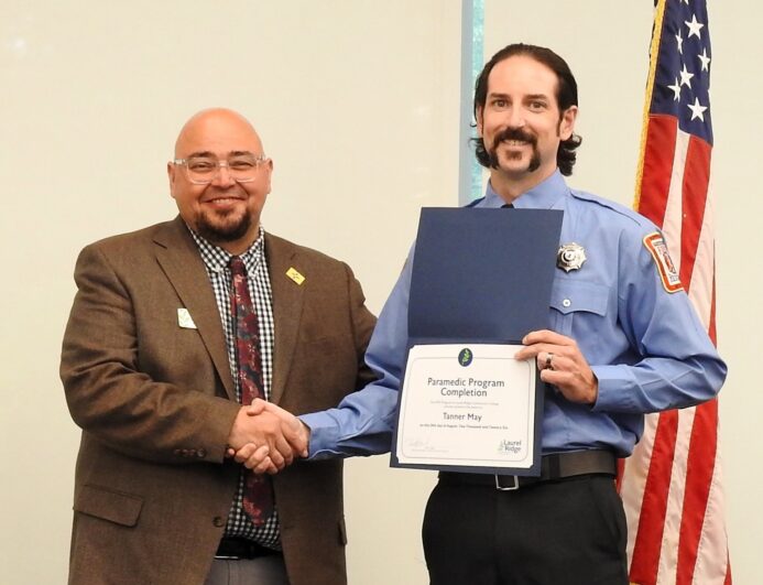 Tanner May shakes hands with a Laurel Ridge Community College representative while holding a certificate for completing the Paramedic Program, with an American flag in the background.