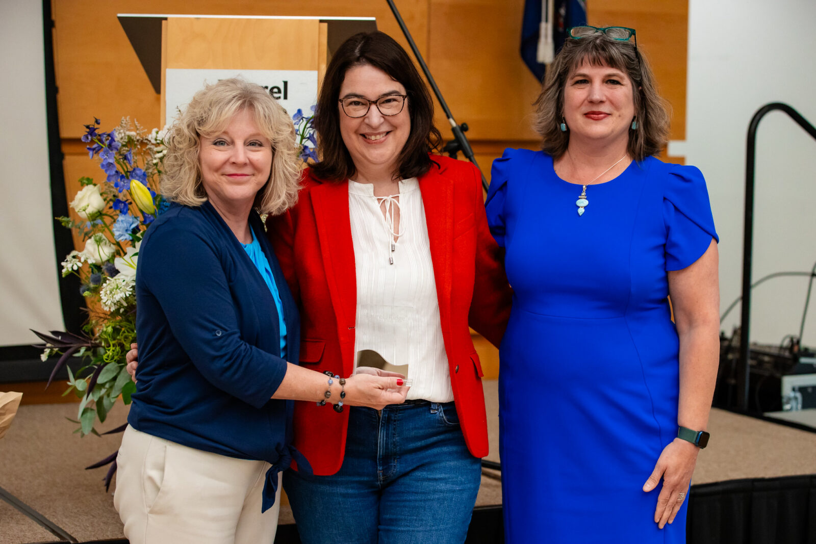 Three women stand smiling together on a stage at a Laurel Ridge Community College event, with one woman in the middle, Nicole Munday, holding a small glass award. She wears a red blazer over a white top with jeans. To her left is a woman with curly blonde hair in a navy top and cream pants, and to her right is a woman with gray-brown hair wearing a royal blue dress and teal earrings. Behind them is a wooden podium with a partial Laurel Ridge sign, a tall floral arrangement with blue and white flowers, and a Virginia state flag.