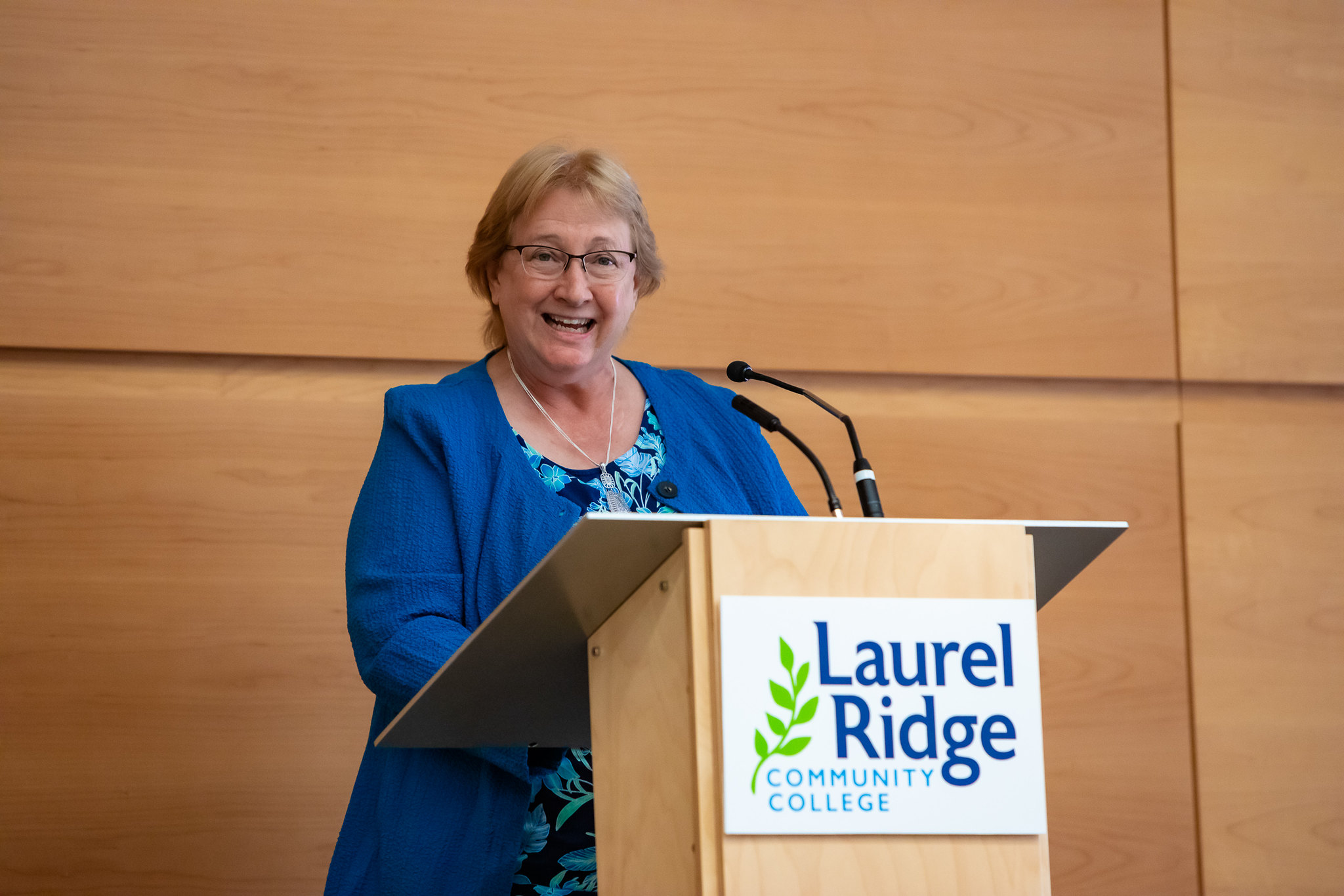 Ann Simpson speaking at a podium with microphones in a wood-paneled room. The podium displays the Laurel Ridge Community College logo, and the speaker is wearing a blue jacket and patterned blue top.
