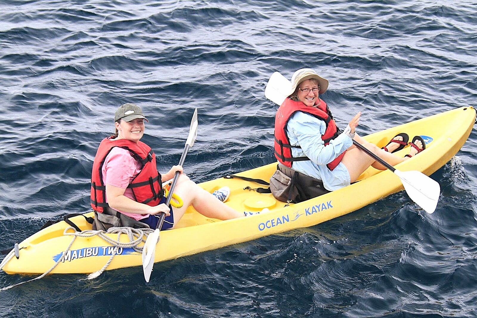 Two people wearing life jackets paddle a yellow tandem kayak on open water, viewed from above. One person waves while both hold double-ended paddles.