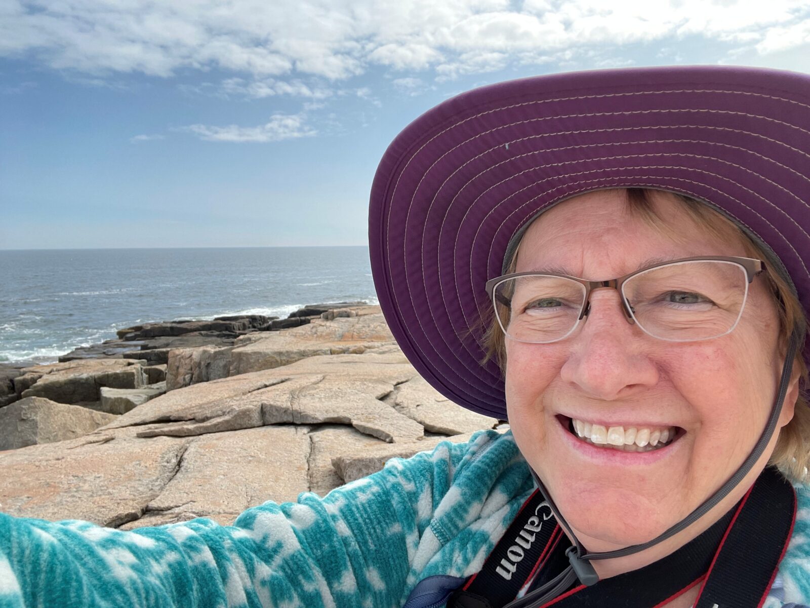 Person taking a selfie on a rocky ocean shoreline, wearing glasses and a wide-brimmed purple sun hat. Waves and the horizon are visible in the background beneath a partly cloudy sky.