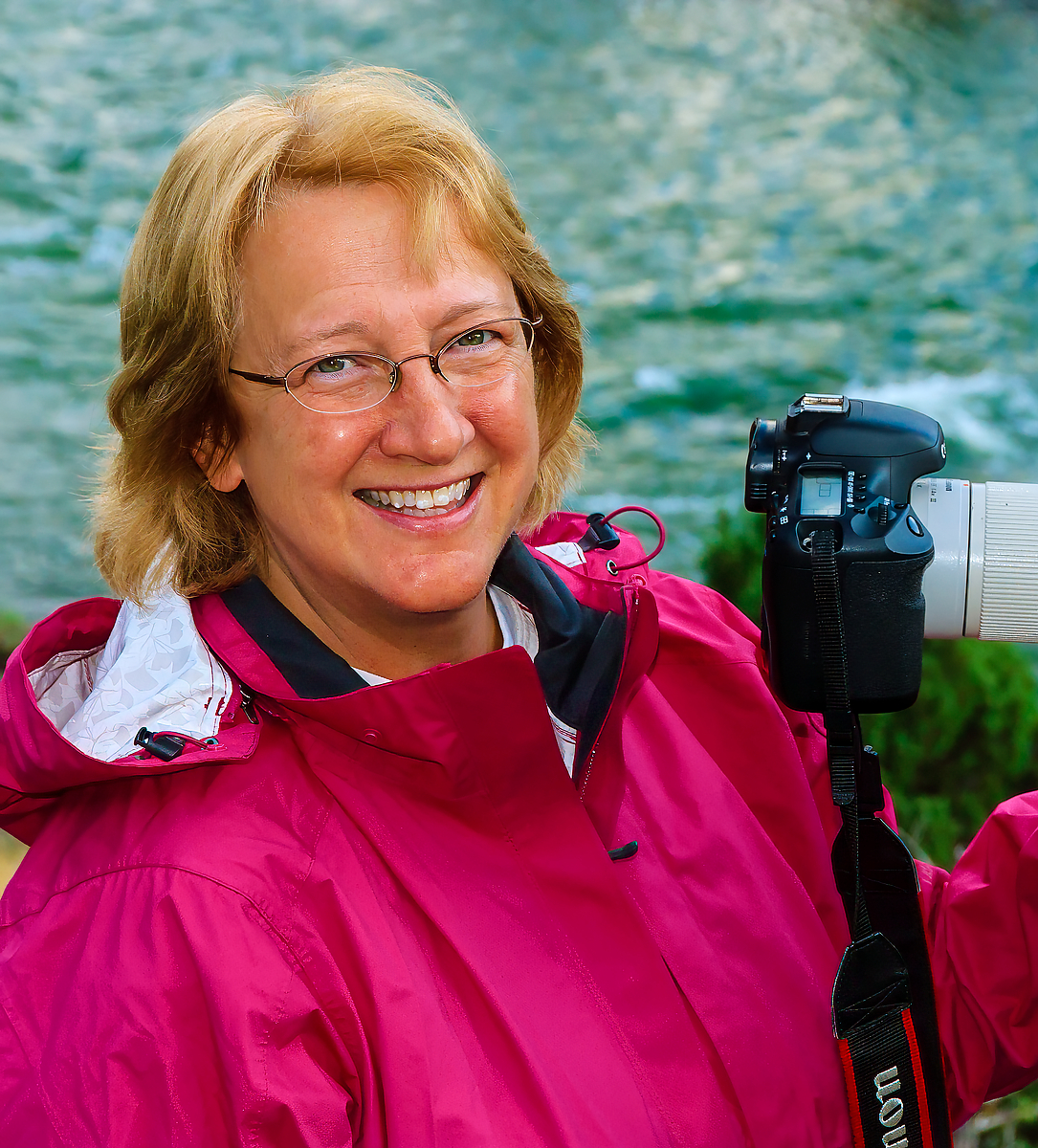 Ann Simpson wearing a rain jacket and holding a camera, taking photos outdoors with a body of water featured behind her.