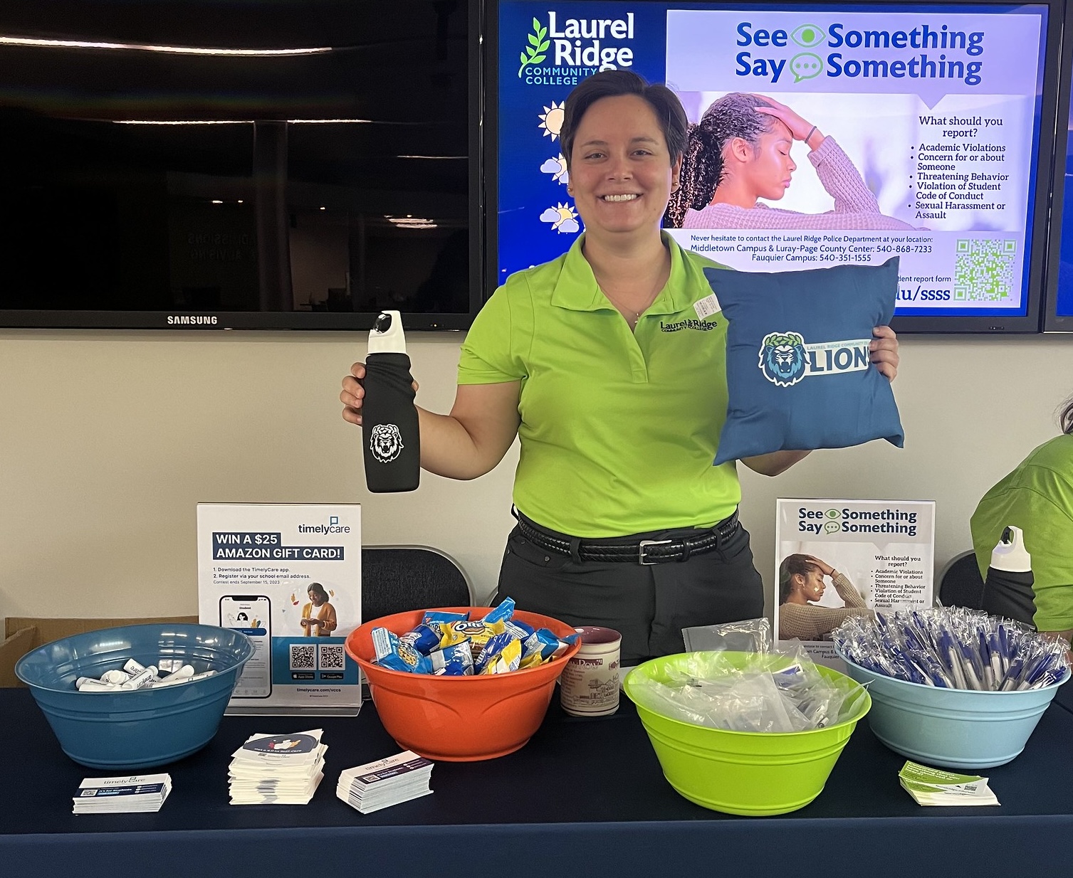 Caroline Walls standing behind an information table, holding a branded water bottle and pillow. The table displays giveaway items, snacks, pens, and resource cards, with digital screens and campus safety information visible in the background.