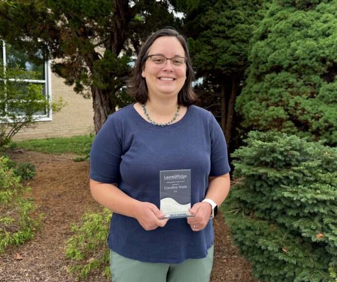Caroline Walls standing outdoors in front of evergreen trees, holding a glass award plaque with both hands. A building and landscaped area are visible in the background.