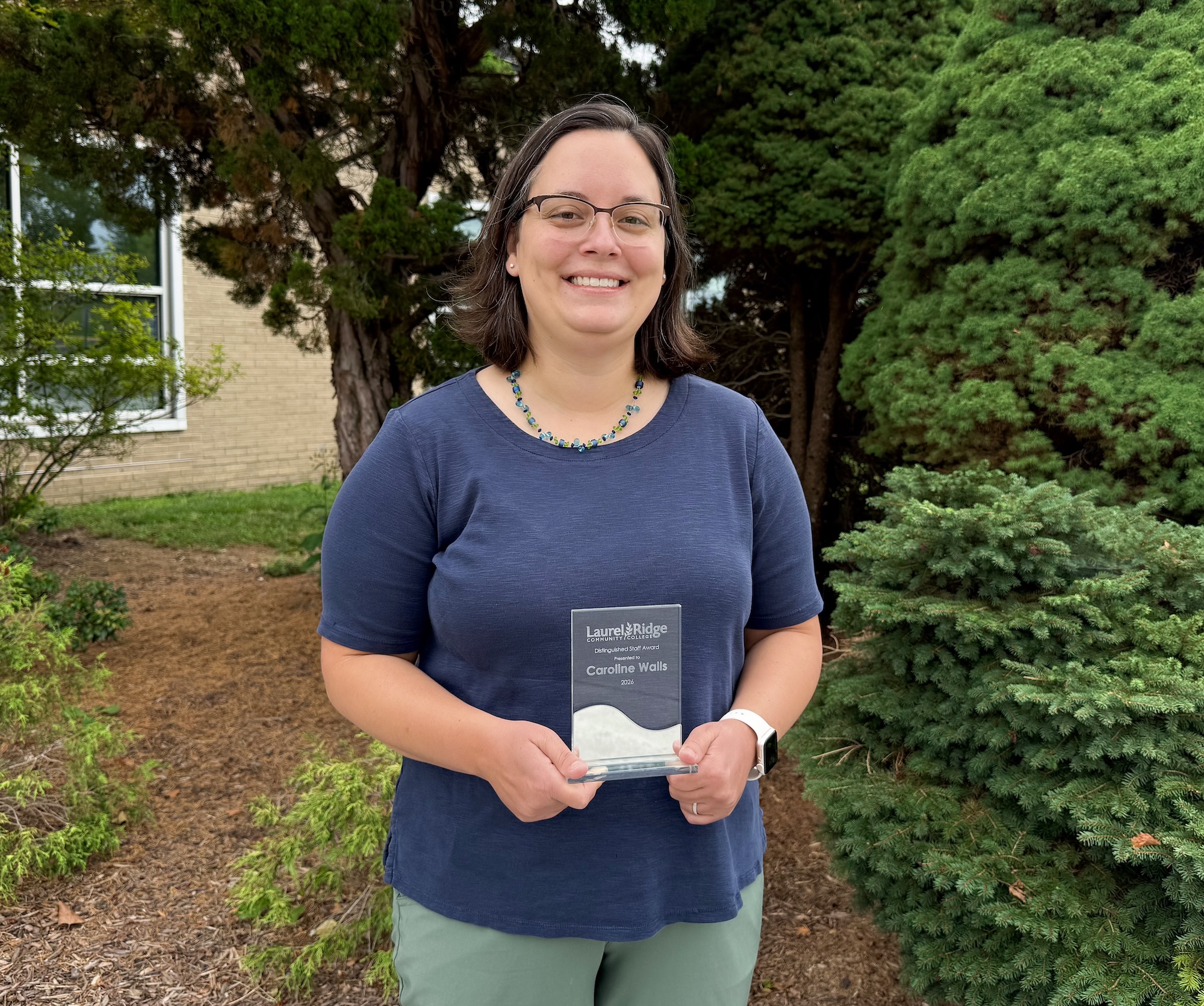 Caroline Walls standing outdoors in front of evergreen trees, holding a glass award plaque with both hands. A building and landscaped area are visible in the background.