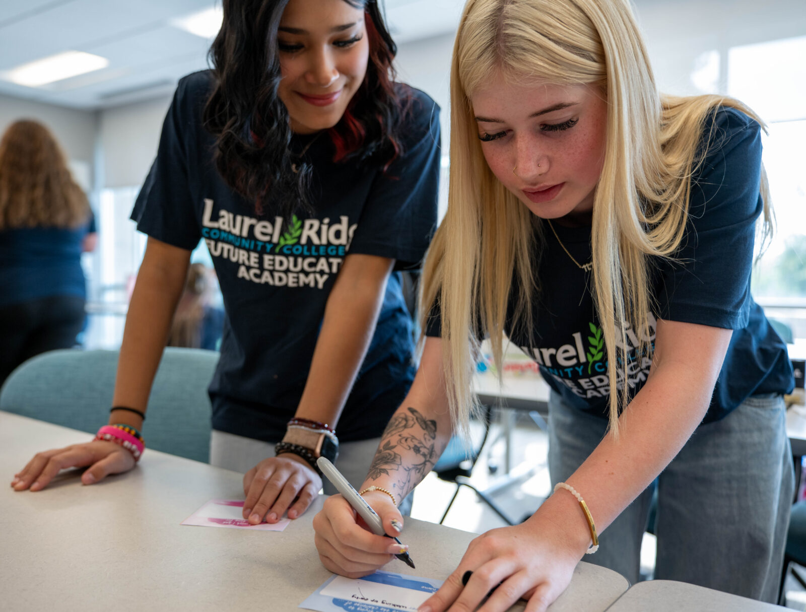 Two Future Educators Academy students collaborate on a classroom activity, with one writing on a card while the other observes. Both wear Laurel Ridge Community College program T-shirts in a bright learning space.