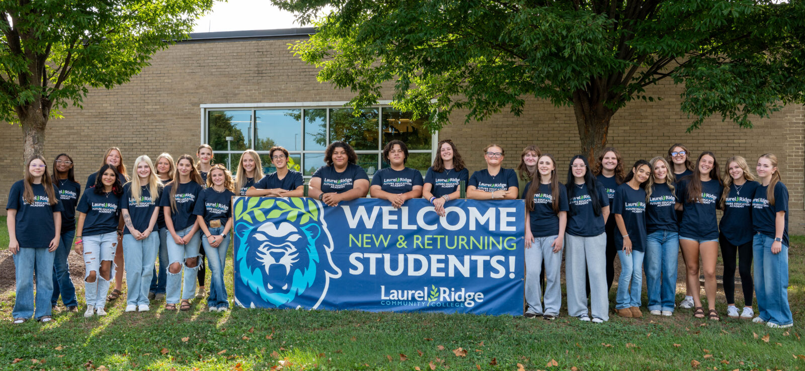 Future Educators Academy students stand outdoors behind a Laurel Ridge Community College welcome banner, posing as a group in matching program T-shirts in front of a campus building and trees.