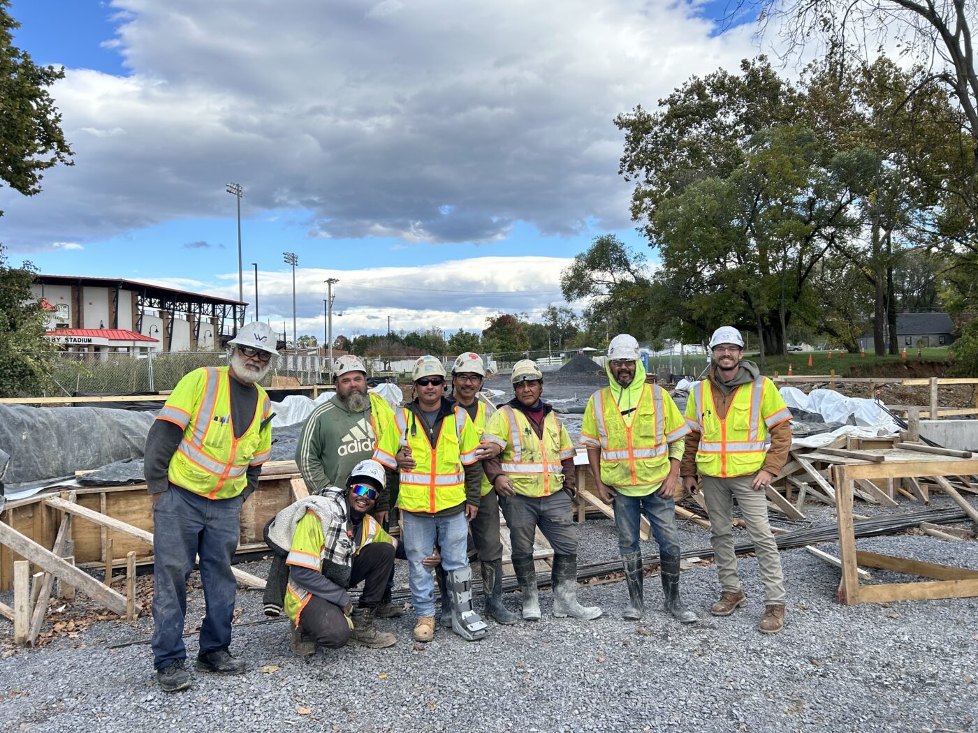 Construction crew wearing high-visibility safety vests and hard hats pose at an active worksite with wood forms, gravel, and building materials. Stadium structures and light poles are visible in the background beneath a dramatic cloudy sky.