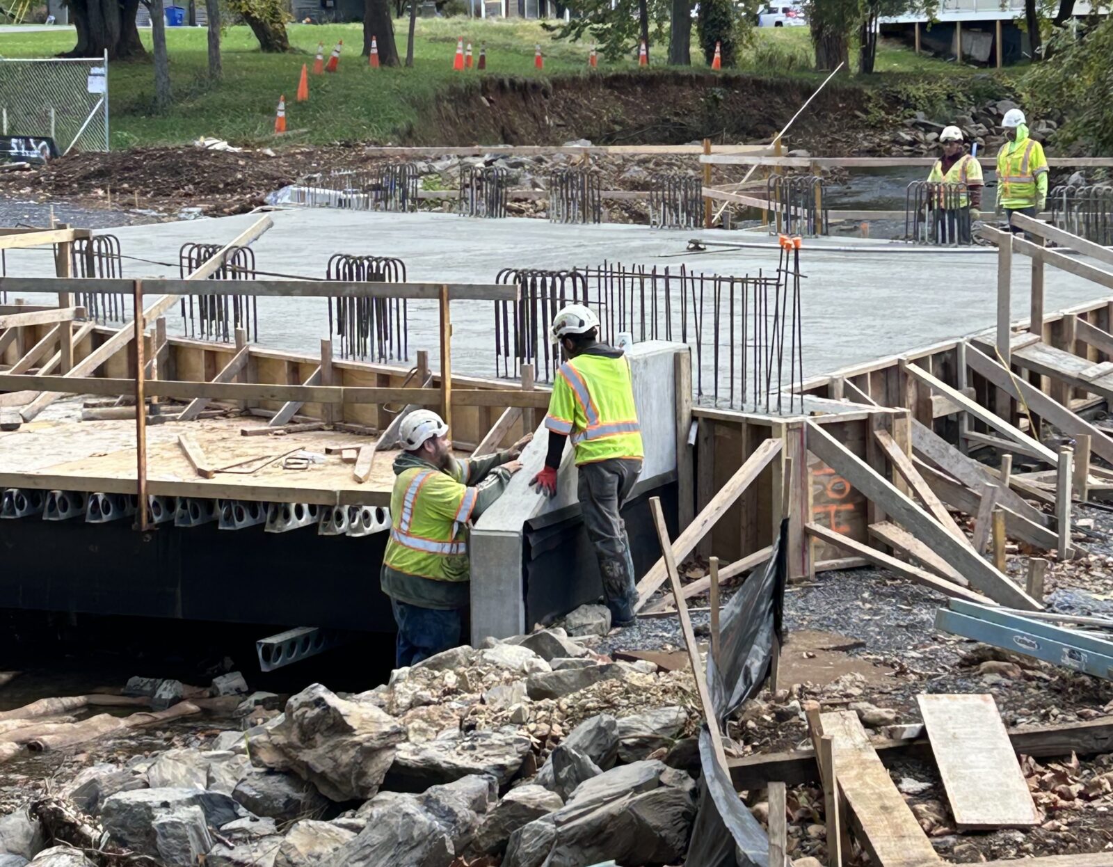 Workers in high-visibility safety gear install concrete components at a bridge or culvert construction site. Freshly poured concrete, exposed rebar, wood formwork, rock stabilization, and active construction activity. 