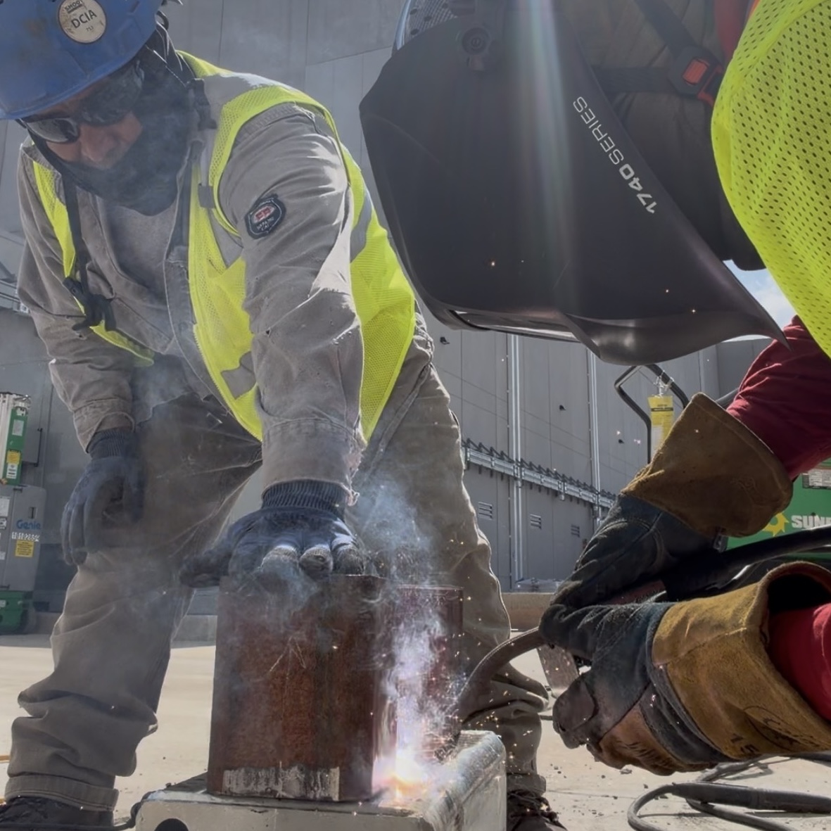 Welding workers wearing safety vests, gloves, and protective gear weld a steel component outdoors, with sparks and smoke visible in front of an industrial building.