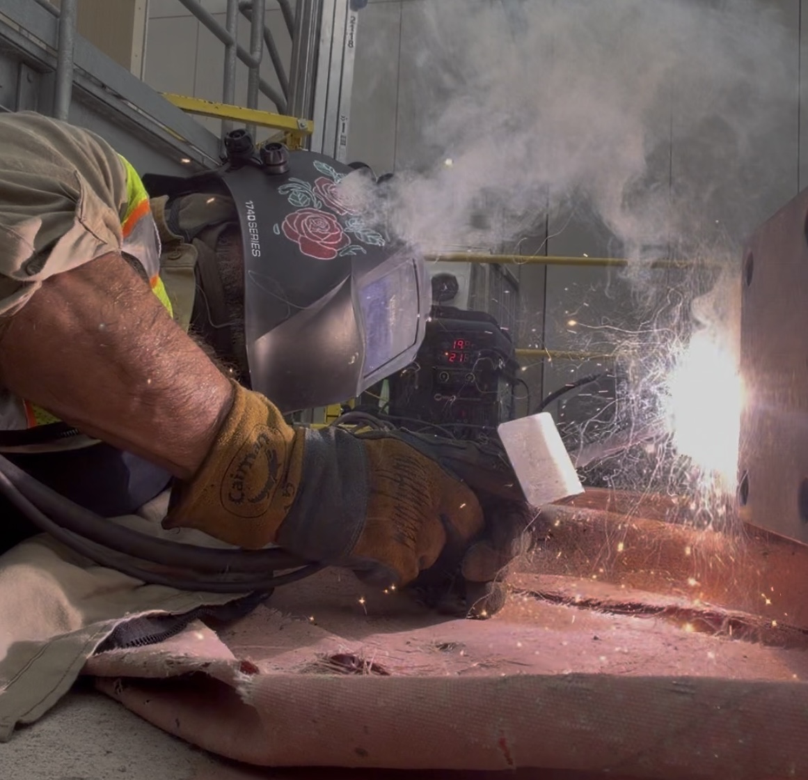 Mike Mullins wearing protective welding gear welds a large metal structure indoors, with bright sparks and smoke visible around the welding torch.