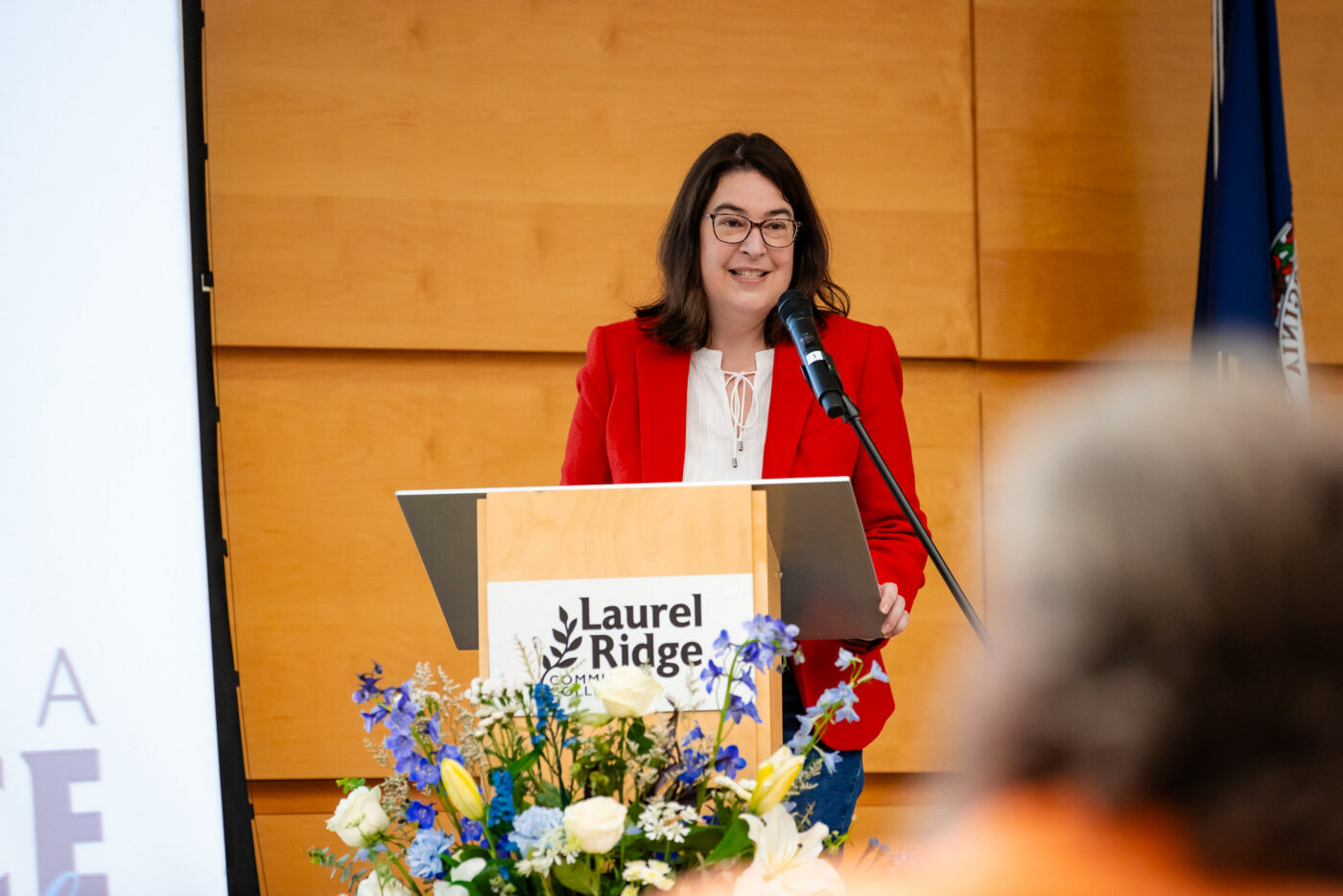 Nicole Munday standing at a podium with a Laurel Ridge Community College sign, speaking into a microphone during an event. A floral arrangement decorates the front of the podium, with audience members visible in the foreground.