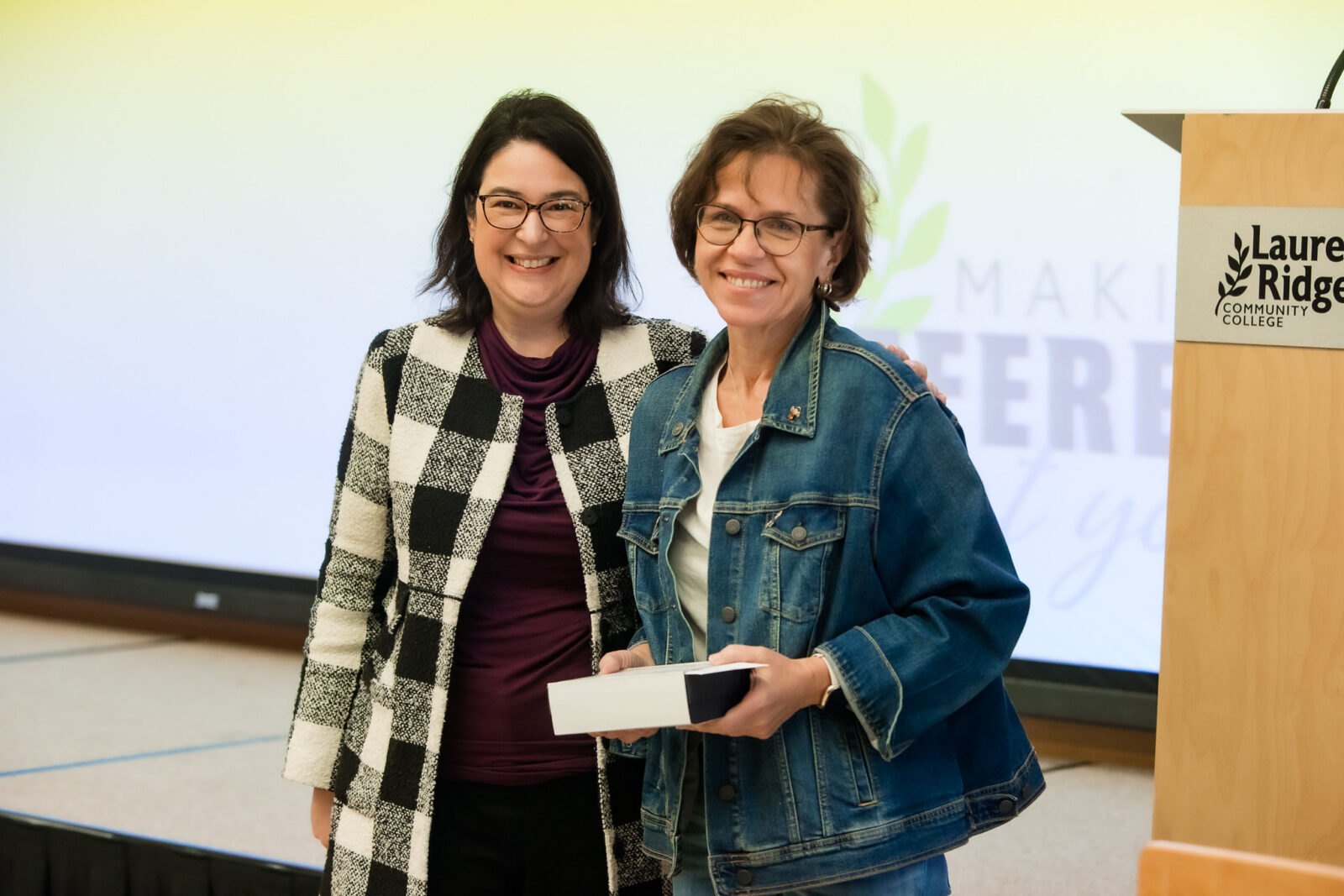 Two people pose together on stage at a Laurel Ridge Community College event. One person holds a small award or presentation box while standing beside the presenter in front of a projection screen.