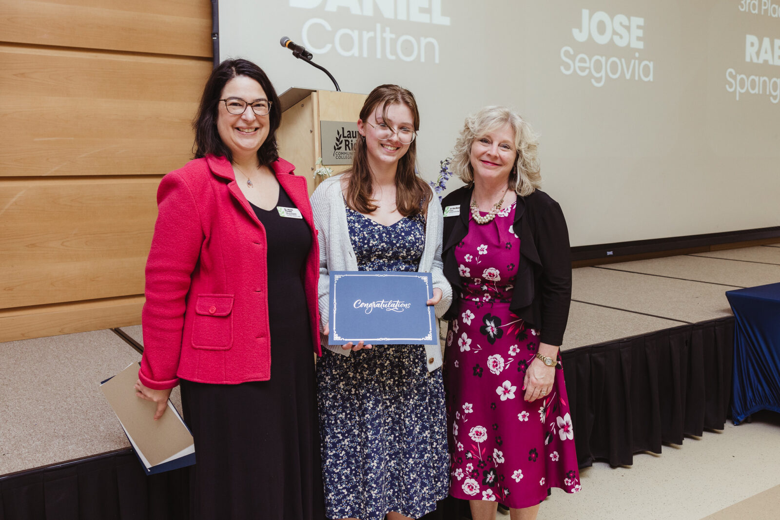 Three people stand together on stage during an awards ceremony at Laurel Ridge Community College. The person in the center holds a certificate while posing with two presenters near a podium and projection screen.