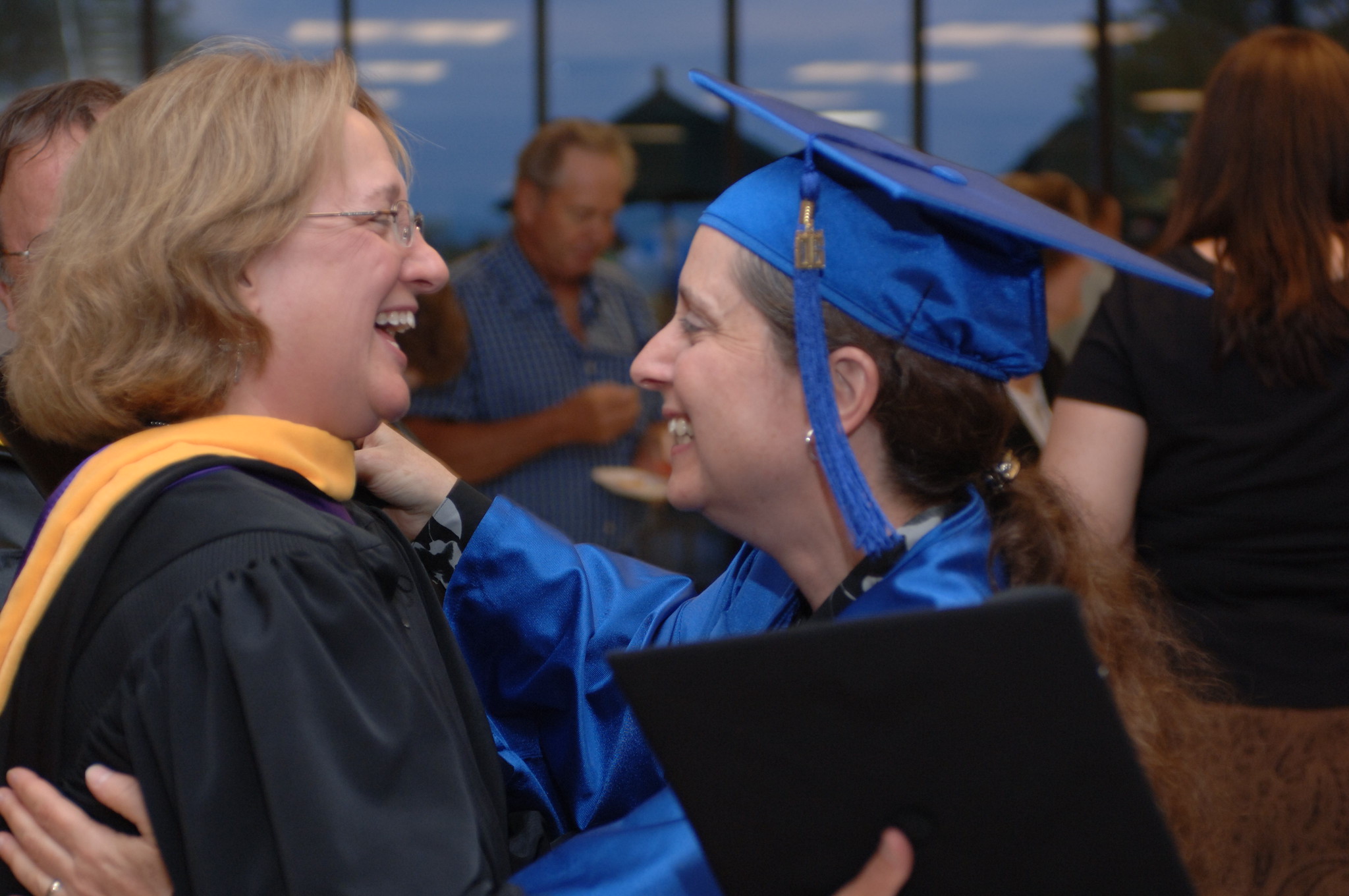 Graduate in a blue cap and gown embraces a faculty member in academic regalia during a commencement celebration, with other attendees blurred in the background.