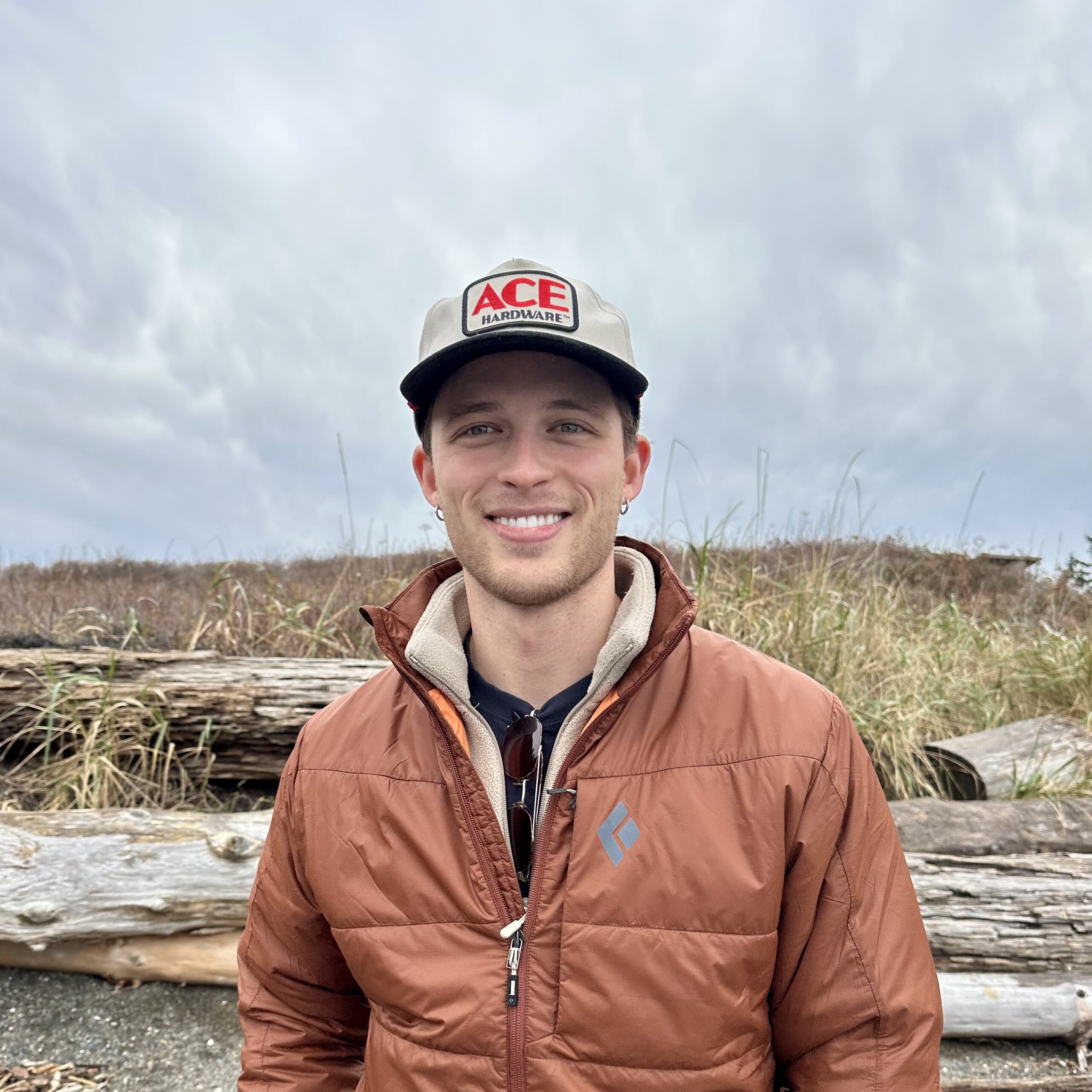 Taylor Bloom wearing a brown insulated jacket and gray ACE Hardware cap standing in front of driftwood logs and tall beach grass under a cloudy sky.