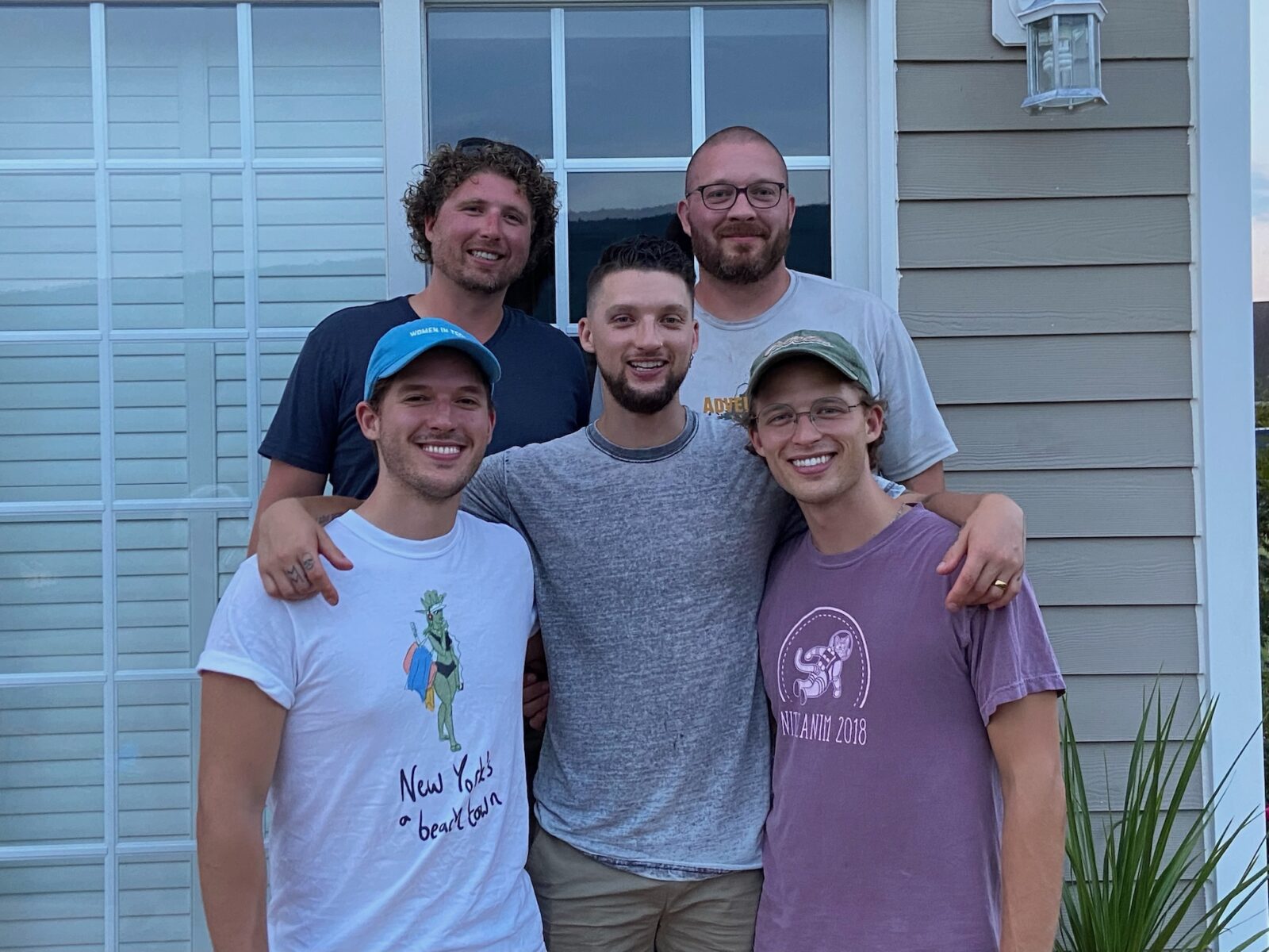 Five people standing close together on a front porch, posing for a group photo in front of large windowed doors and house siding. Two people stand in back and three in front, with arms around each other. Casual clothing and baseball caps; a plant is visible at right.