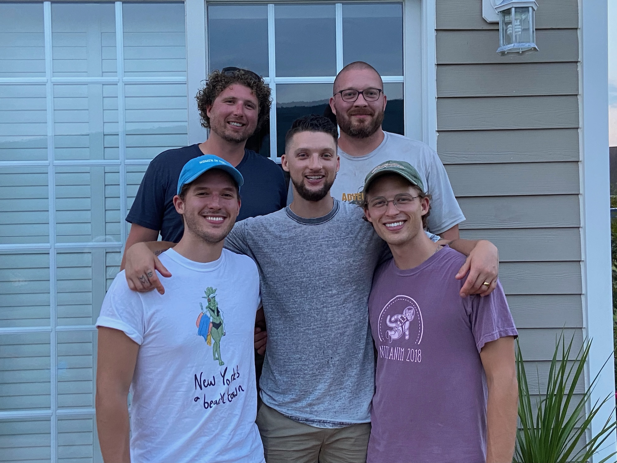 Five people standing close together on a front porch, posing for a group photo in front of large windowed doors and house siding. Two people stand in back and three in front, with arms around each other. Casual clothing and baseball caps; a plant is visible at right.