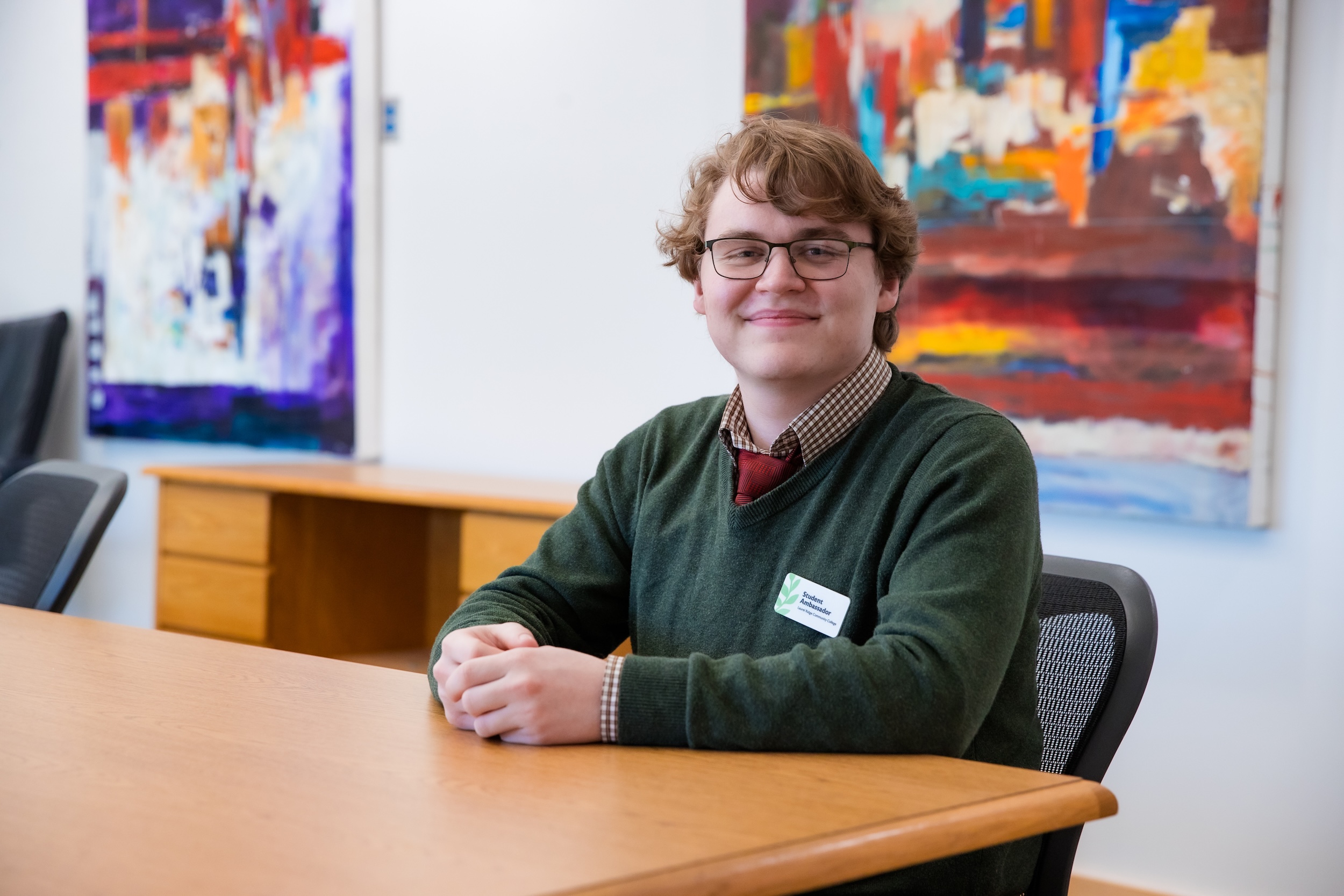 Adam Gagnon, wearing a Student Ambassador name tag, smiles while seated at a table in front of colorful abstract paintings.