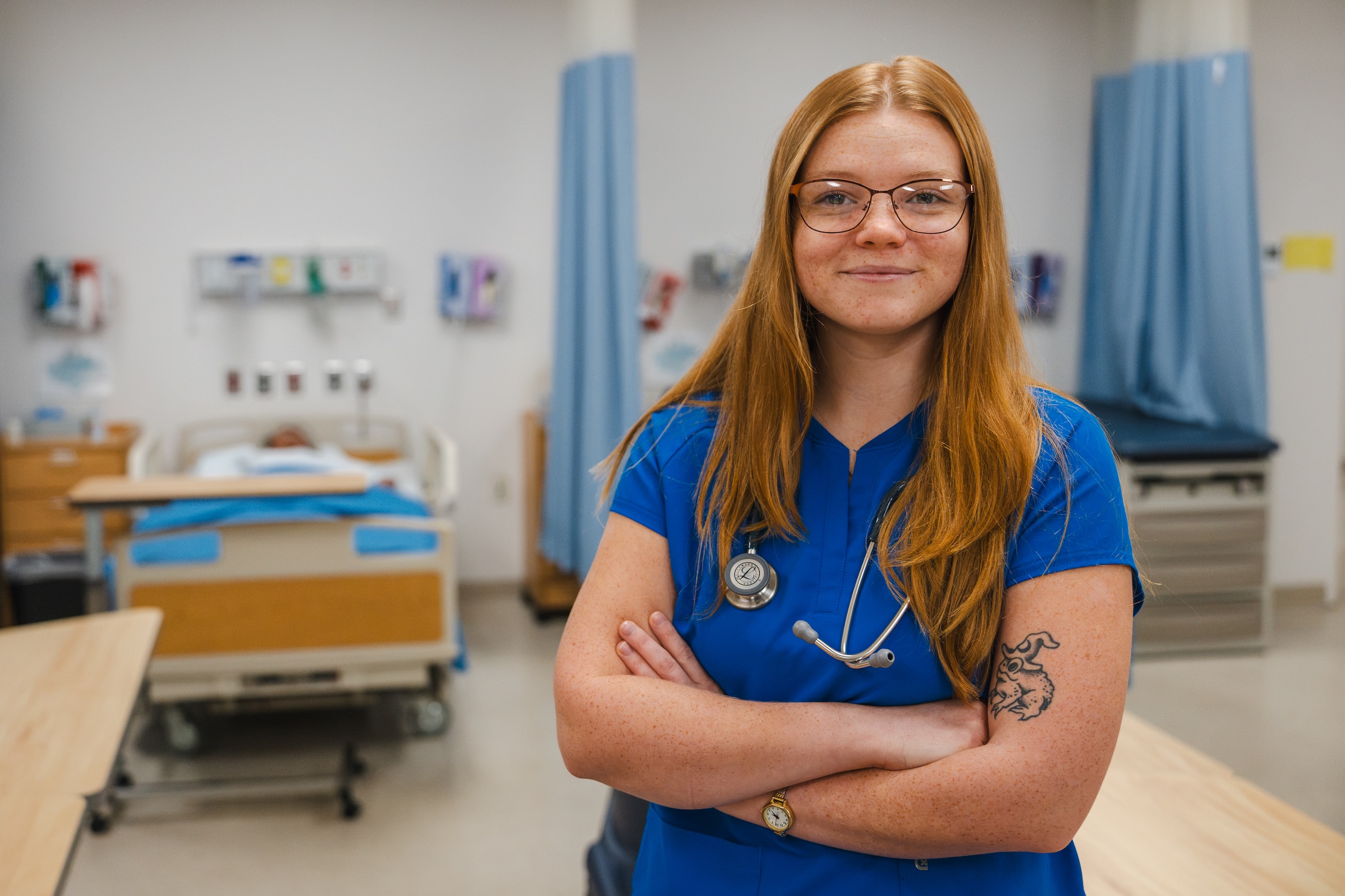 Laila Hall, in blue scrubs with a stethoscope, stands with arms crossed in a Laurel Ridge nursing lab.