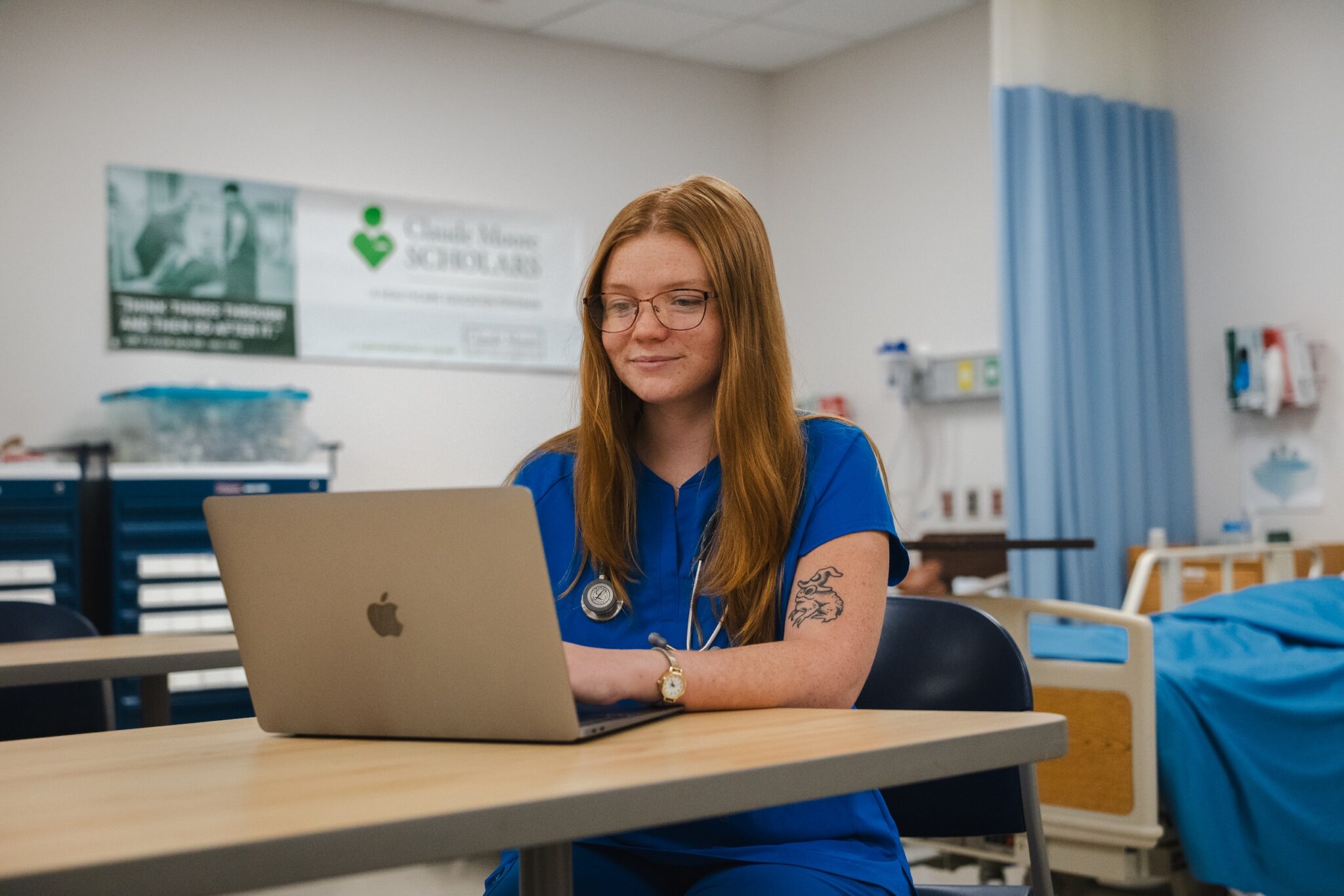 Laila Hall, in blue scrubs with a stethoscope, works on a laptop at a table in a Laurel Ridge nursing lab.