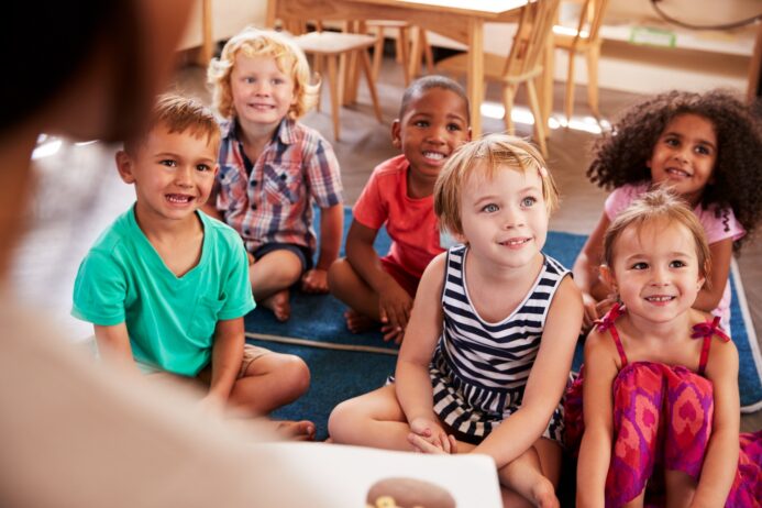 Seven smiling young children sit cross-legged on a classroom rug, looking up at a teacher who is reading to them.