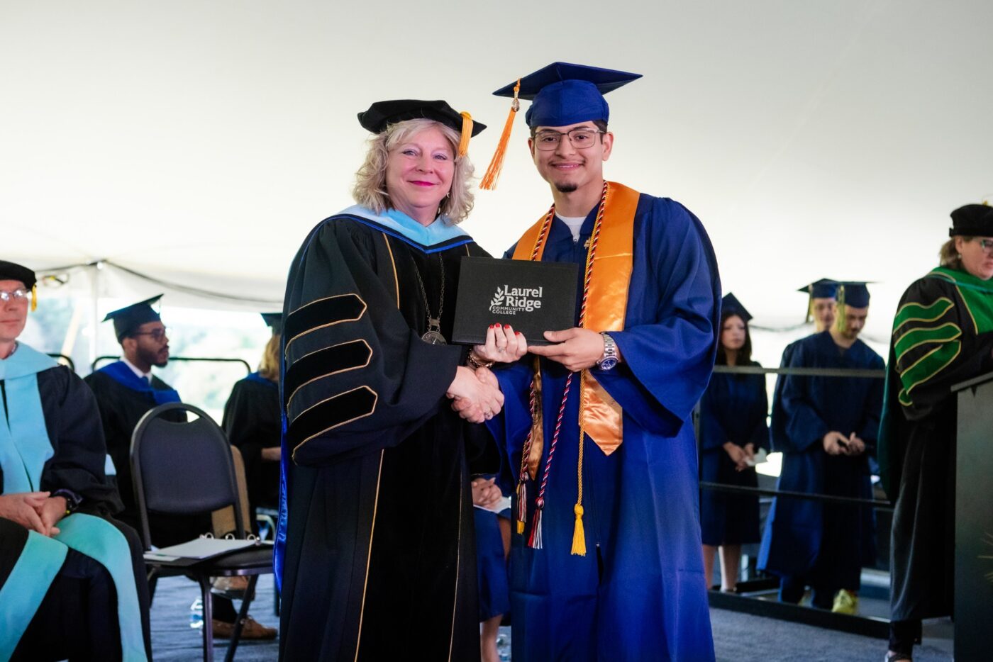 Emanuel Ayala, in a blue cap and gown with an orange stole and honor cords, shakes hands with a college official in doctoral regalia as he receives his diploma cover at commencement.