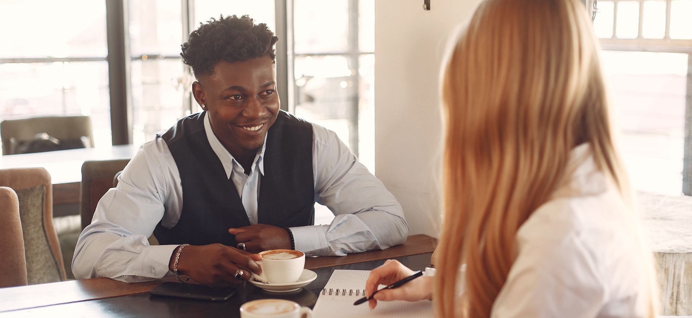 A smiling young man in a vest and dress shirt talks with a woman taking notes over cups of coffee at a café table.