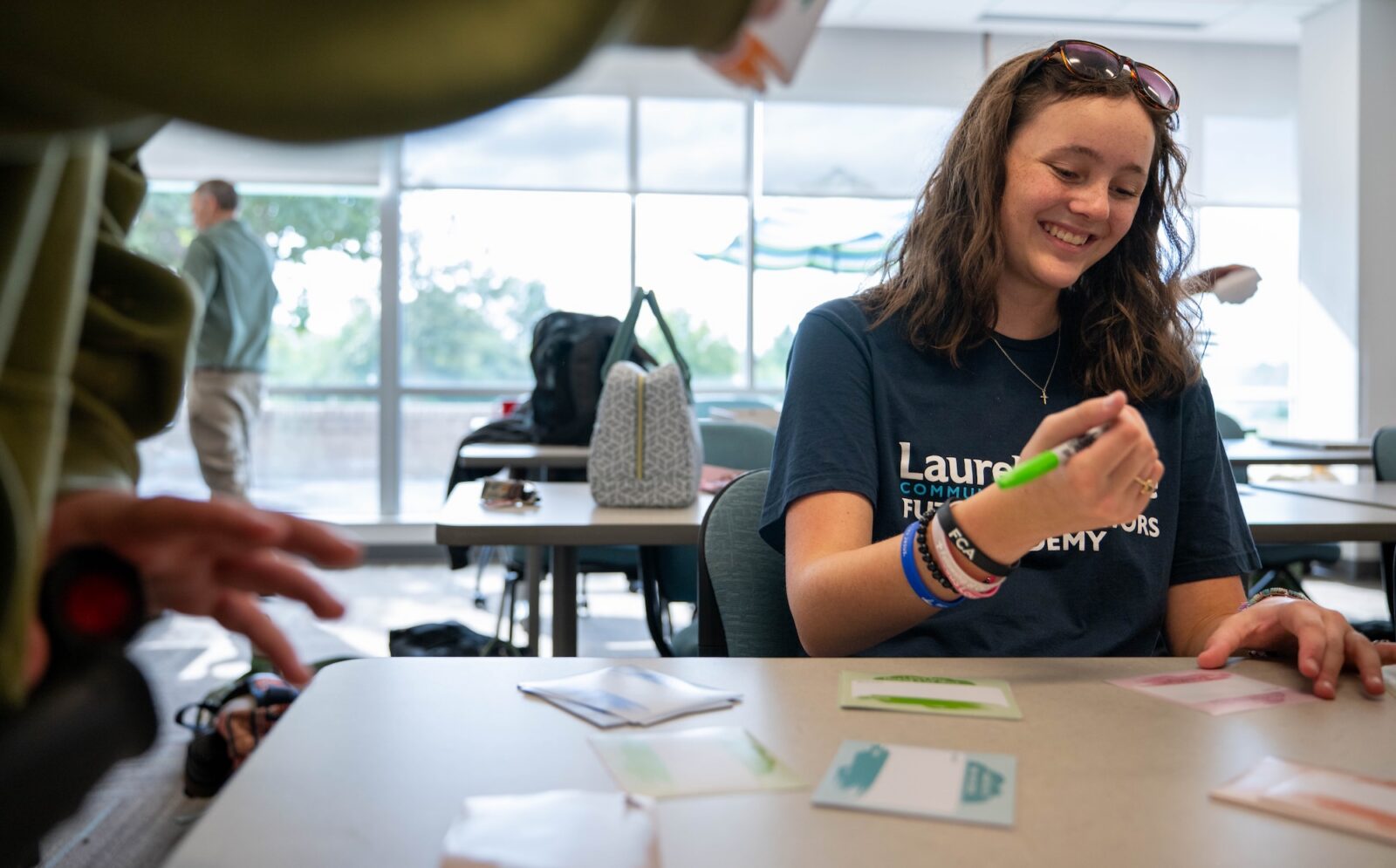 Smiling student in a Laurel Ridge Community College Future Educators Academy t-shirt holding a green marker at a classroom table with colorful cards