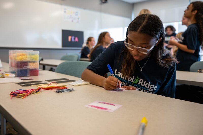 Student in a Laurel Ridge Community College Future Educators t-shirt writing on a card with a blue marker at a classroom table with craft supplies