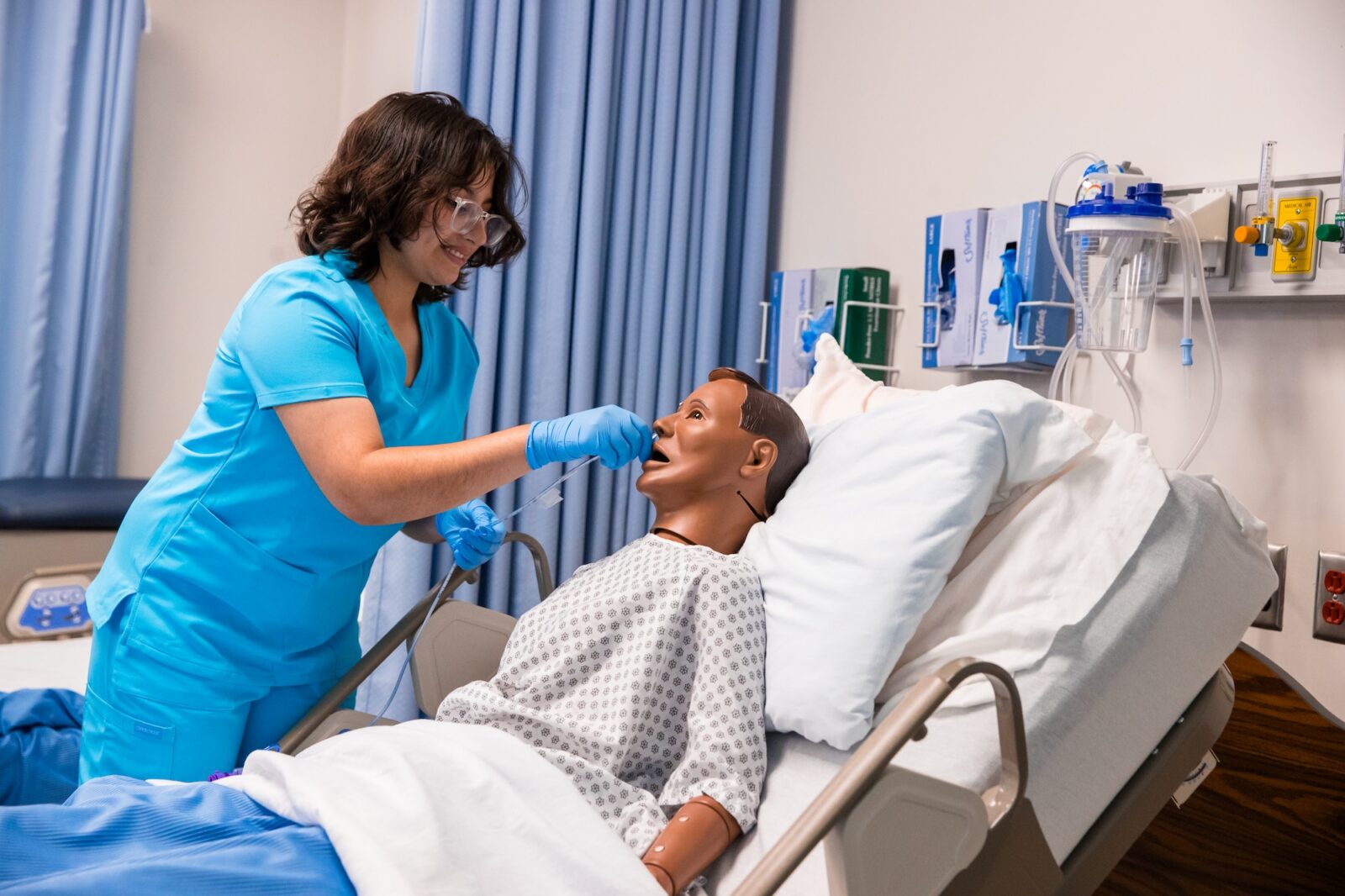 Person in blue scrubs using a medical swab on a patient simulator in a healthcare training room.