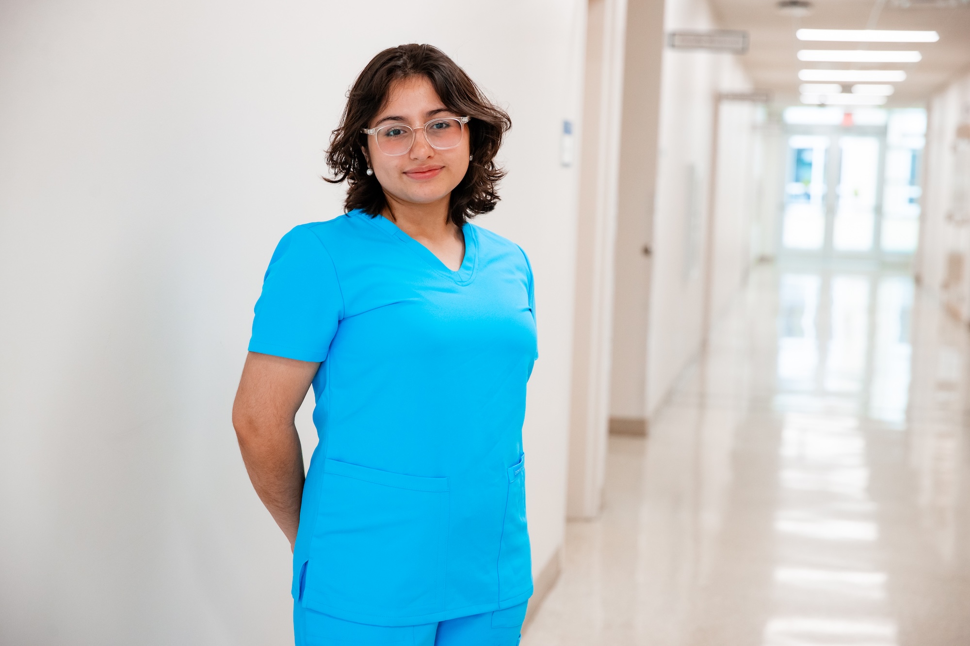 Person wearing bright blue medical scrubs and glasses standing in a healthcare facility hallway.