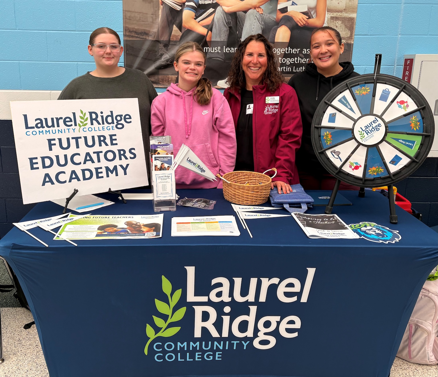 Four people smiling behind a Laurel Ridge Community College Future Educators Academy information table with brochures, pennants and a prize wheel