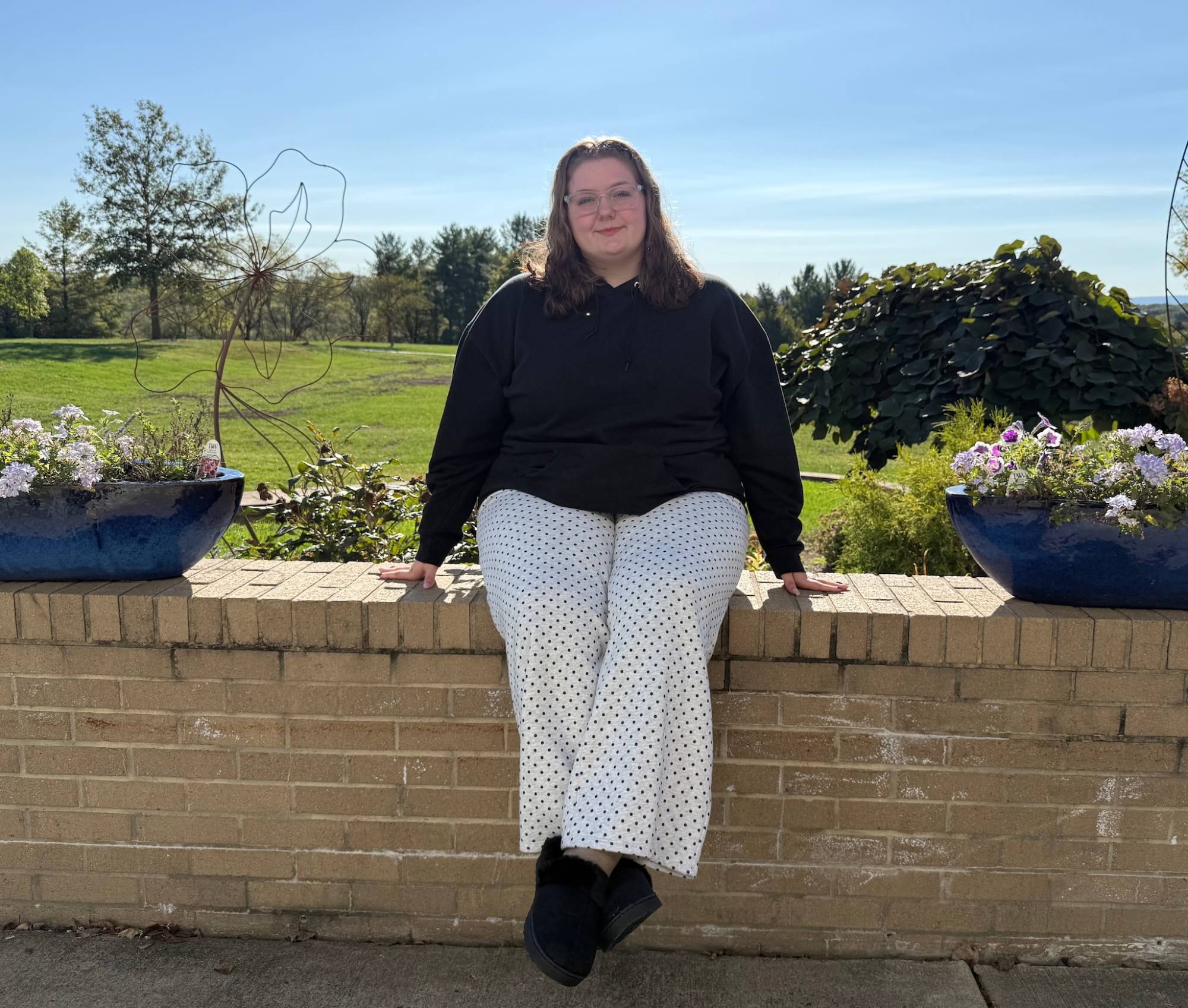Skyla Stewart sits on a brick wall between flower planters with green fields behind her