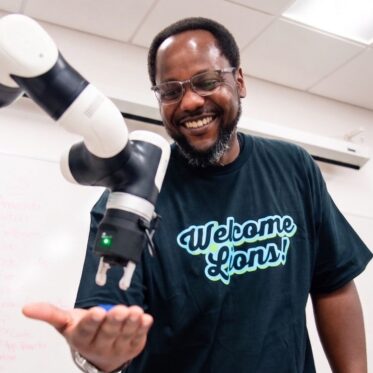 Smiling man in a Welcome Lions T-shirt holding out his hand as a robotic arm's gripper reaches toward it