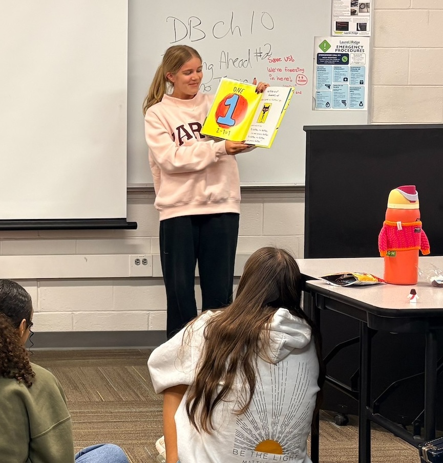 Student standing at the front of a classroom reading a children's picture book aloud to classmates seated on the floor
