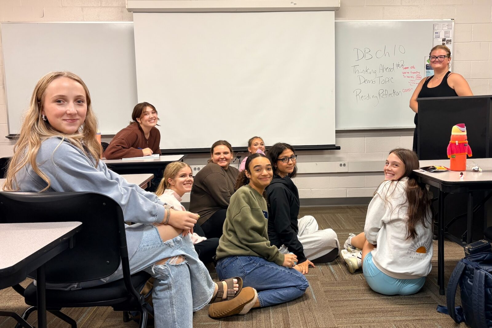 Group of students smiling in a classroom, some seated on the floor and others at desks, with an instructor standing at a podium beside a whiteboard